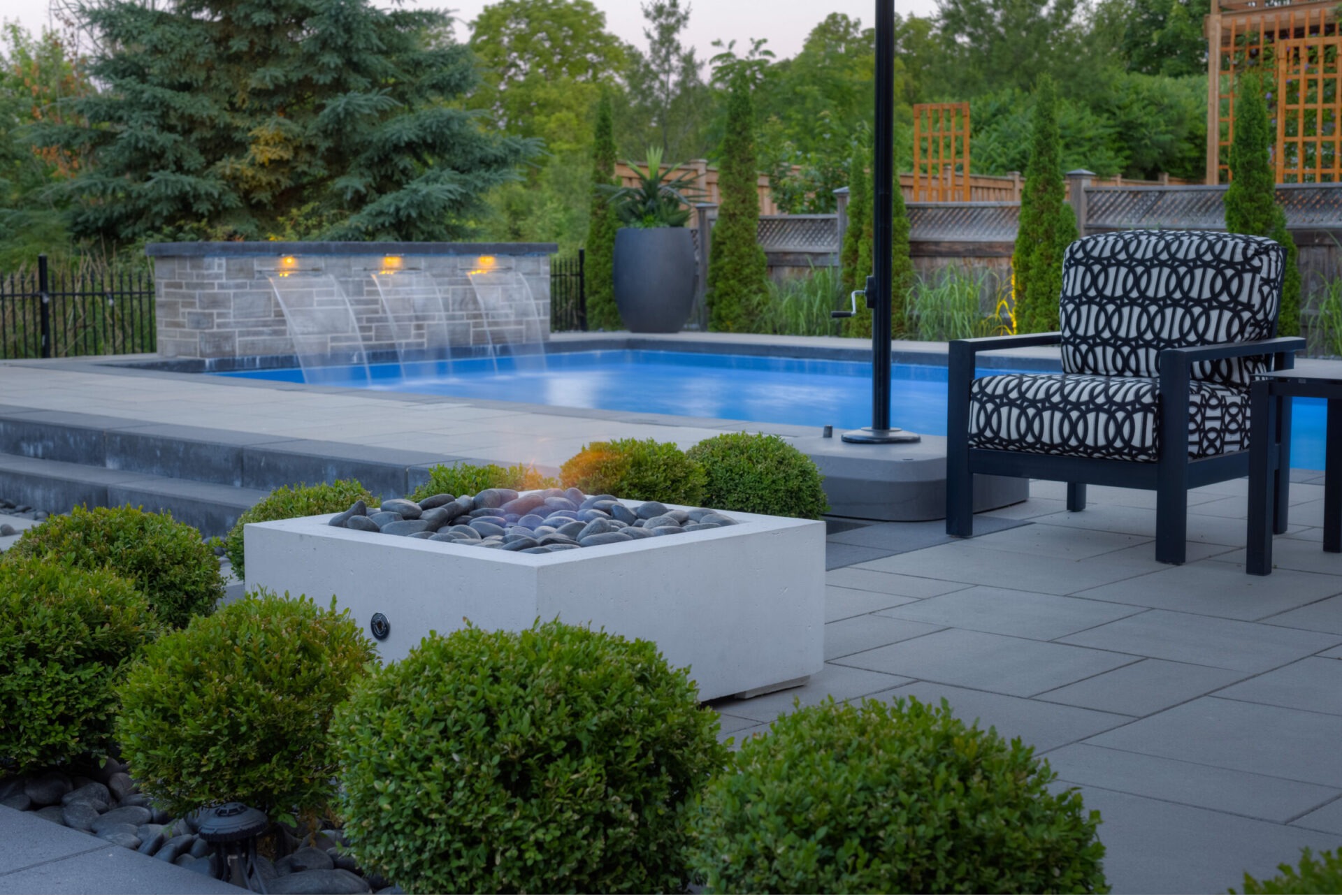 An outdoor pool area during dusk with a lit fire feature, patterned armchair, neatly landscaped bushes, and water flowing from stone pool walls.