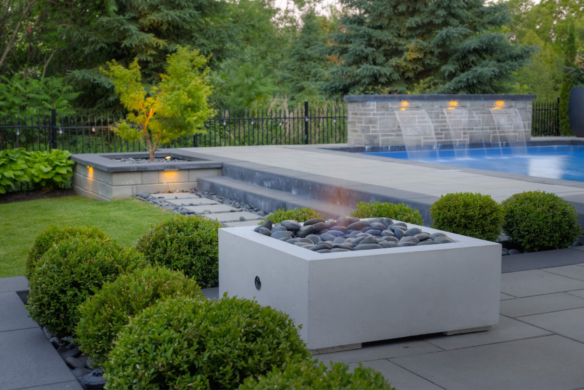An outdoor area with a rectangular fire pit, neatly trimmed hedges, and a swimming pool with cascading water features in the background.