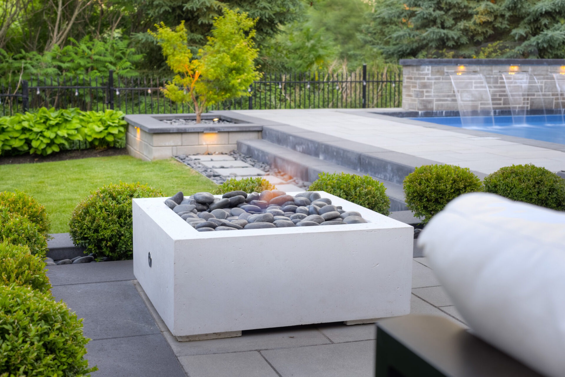 An outdoor space featuring a square fire pit with stones, trimmed bushes, an inviting swimming pool with fountains, and comfortable seating areas.