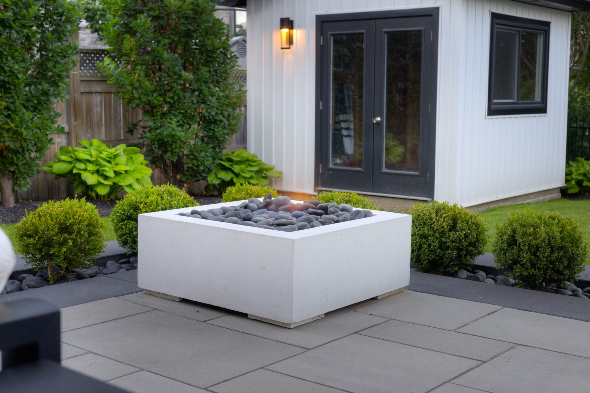 A modern backyard with a square concrete fire pit filled with stones, surrounded by manicured bushes, against a backdrop of a white house.