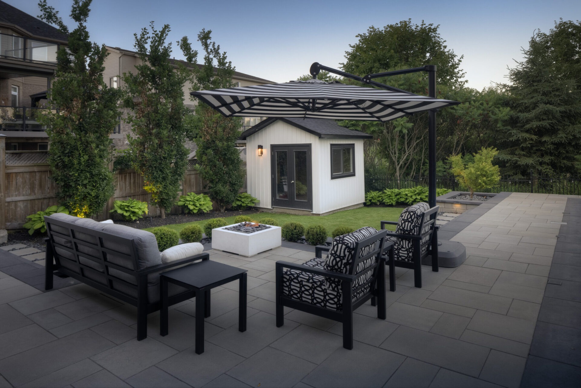 This image features a modern outdoor patio with sleek furniture, a fire pit, a pergola, manicured landscaping, and a small white structure in the background.