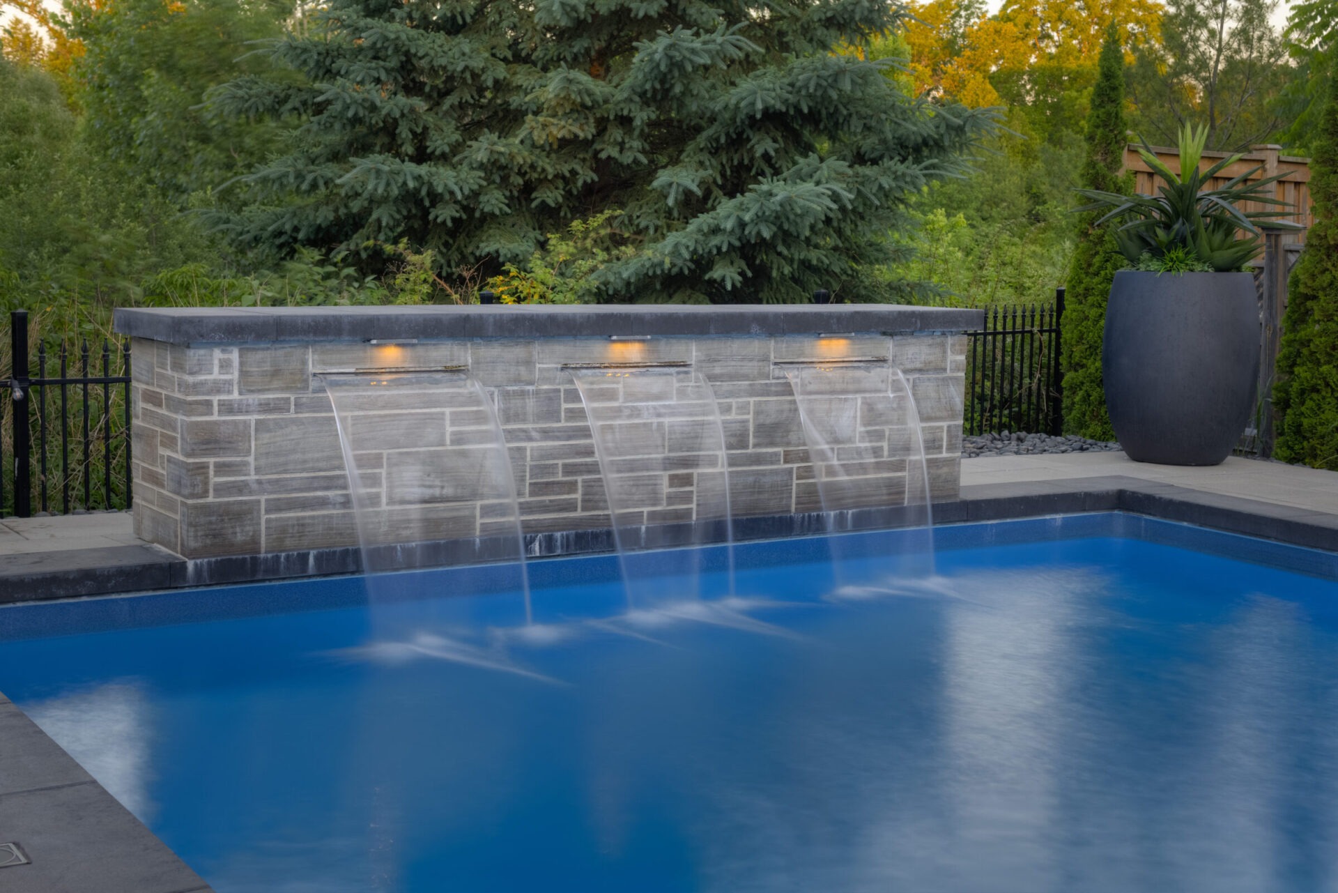 An outdoor swimming pool with a stone wall featuring water cascades, surrounded by lush greenery and a large decorative pot, at dusk.