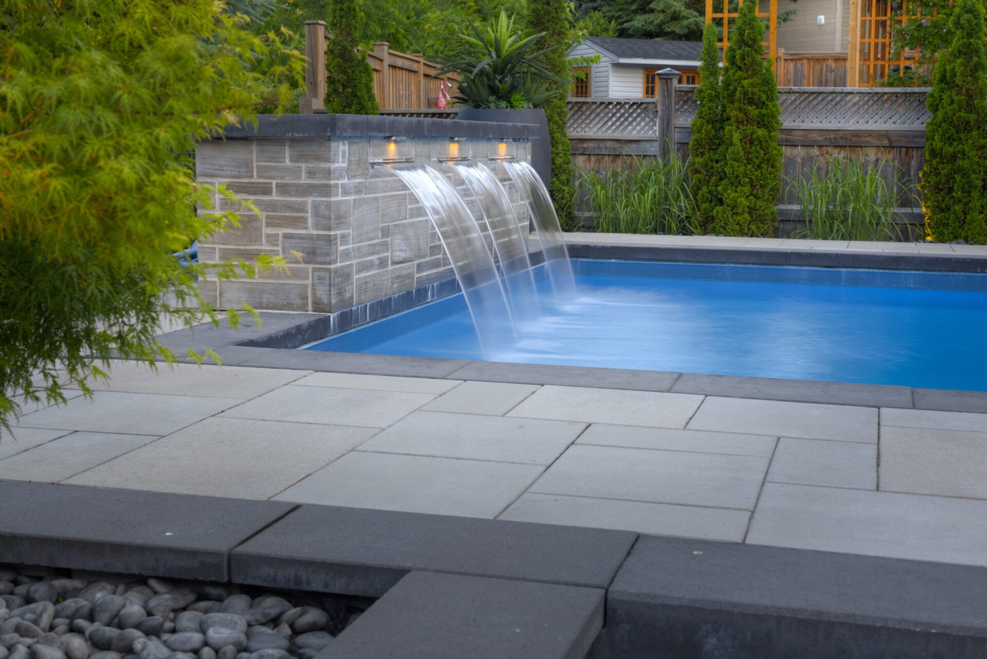 An elegantly landscaped backyard with a pool featuring waterfalls and lights. Paved stones and decorative rocks outline the tranquil space during dusk.