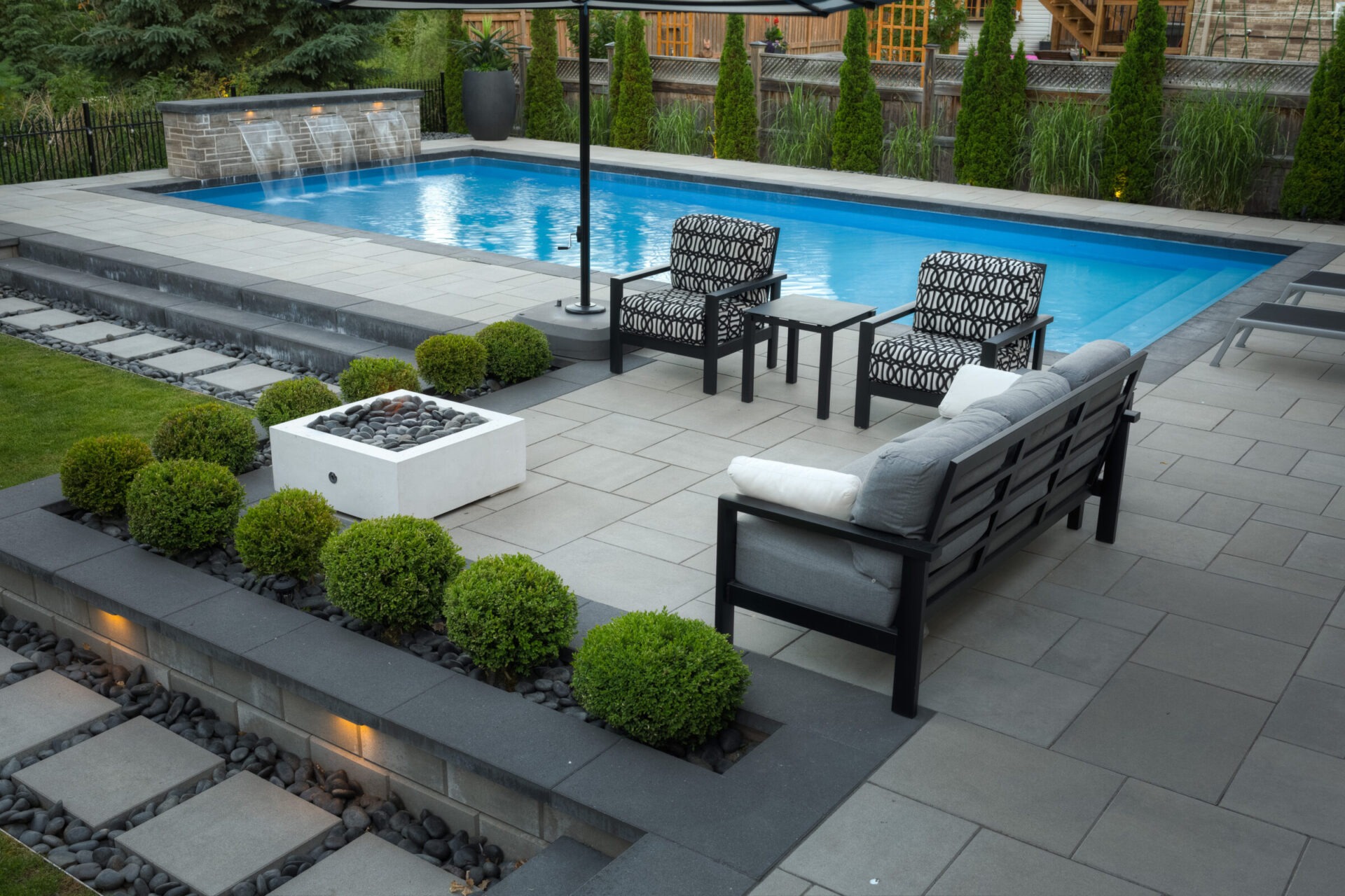 This image shows an elegant backyard with a swimming pool, patterned patio furniture, neat shrubbery, a fire pit, and ambient lighting at dusk.