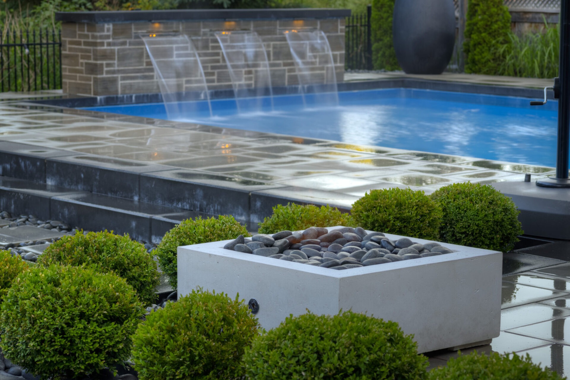 A serene outdoor area featuring a blue swimming pool with water fountains, surrounded by lush green shrubs and smooth grey stones in foreground.