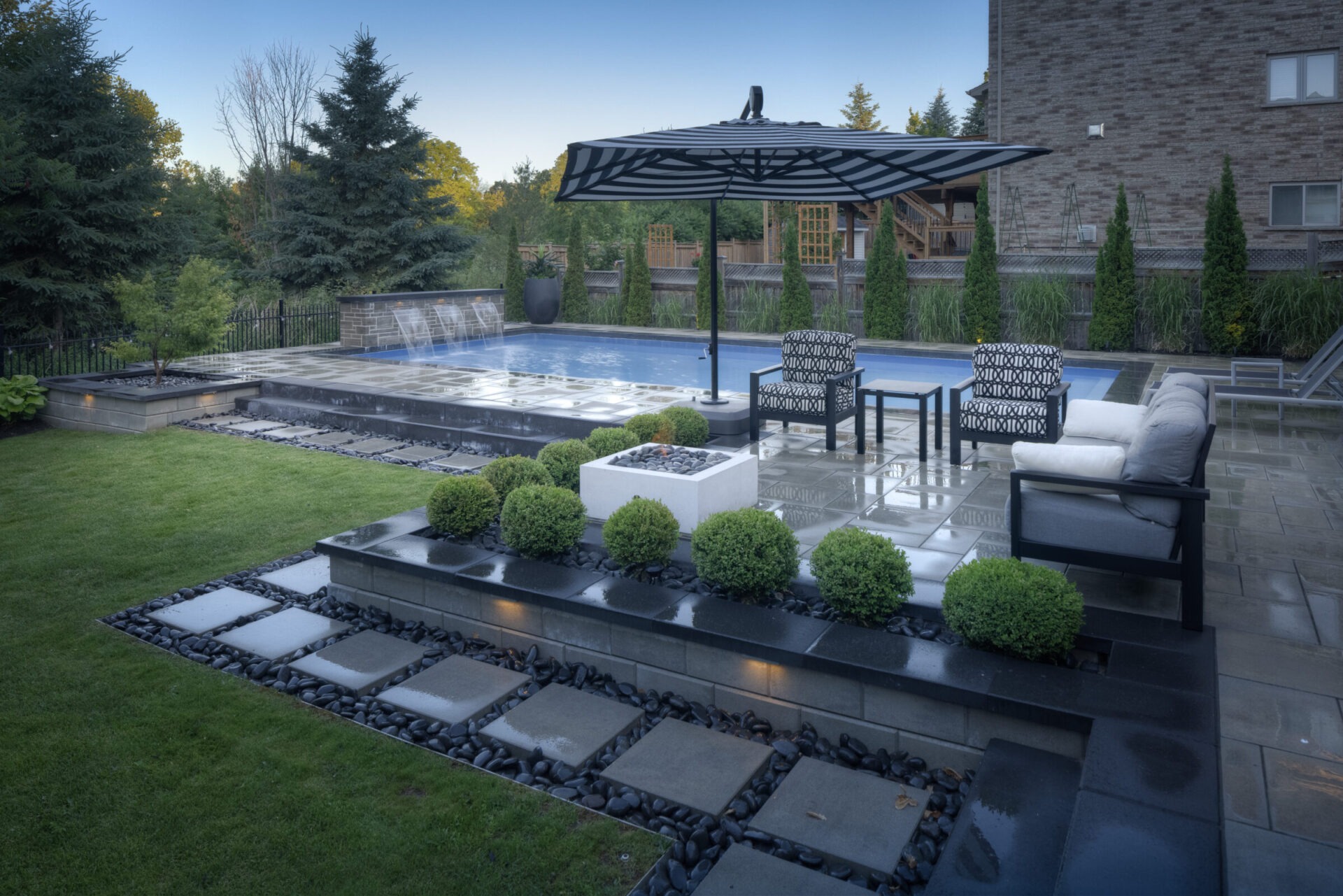 An elegant outdoor space featuring a swimming pool, stylish furniture under a parasol, patterned tile flooring, manicured bushes, and a backdrop of trees.