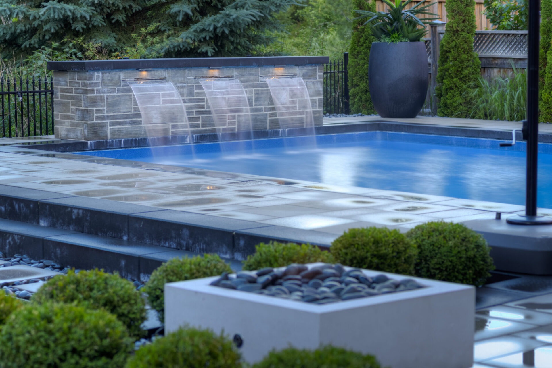 This image displays an elegant outdoor swimming pool with water features streaming down, surrounded by neatly landscaped plants and decorative lighting.