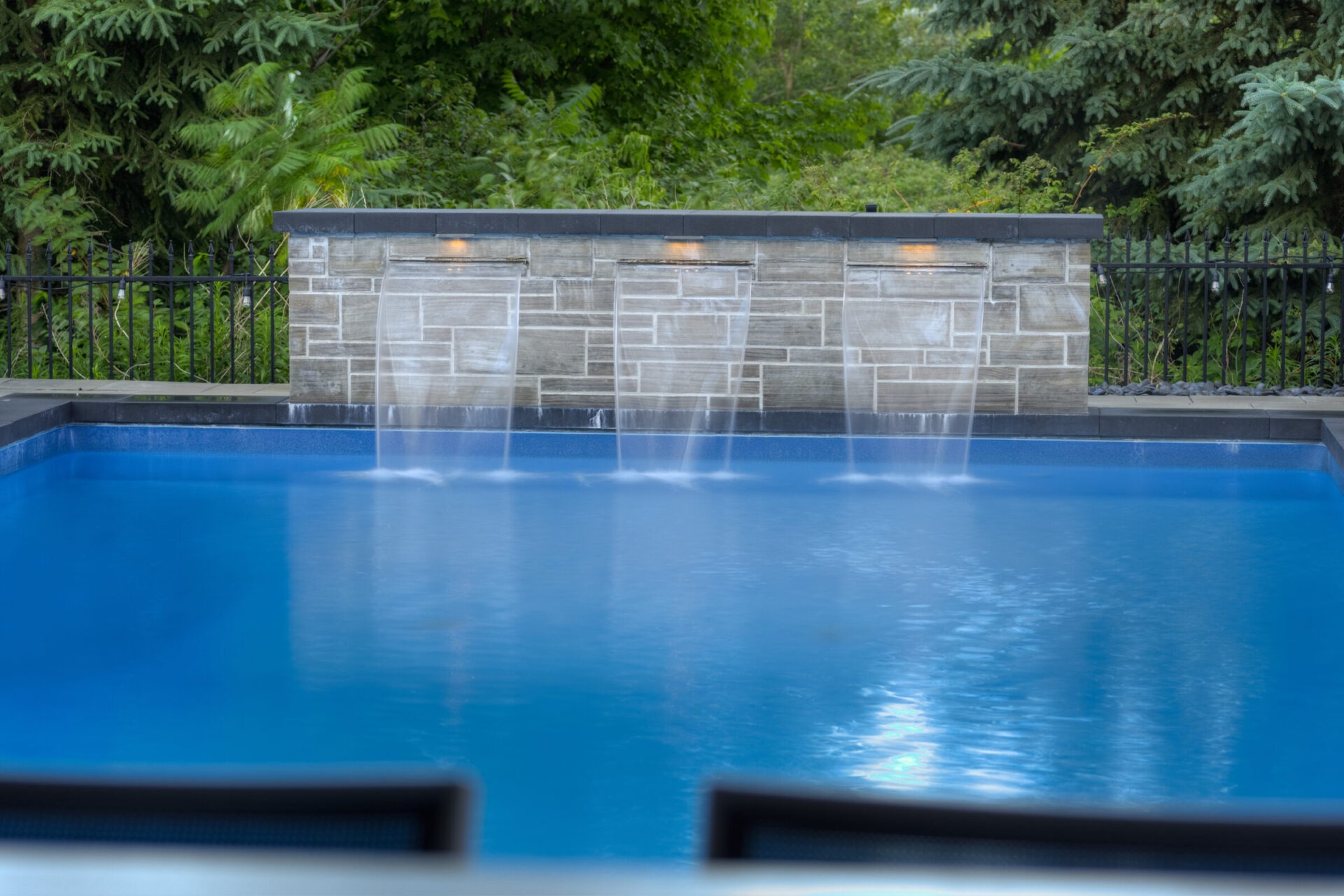A serene swimming pool with tranquil water features pouring into the blue water. Surrounded by green foliage and a stone wall with integrated lights.