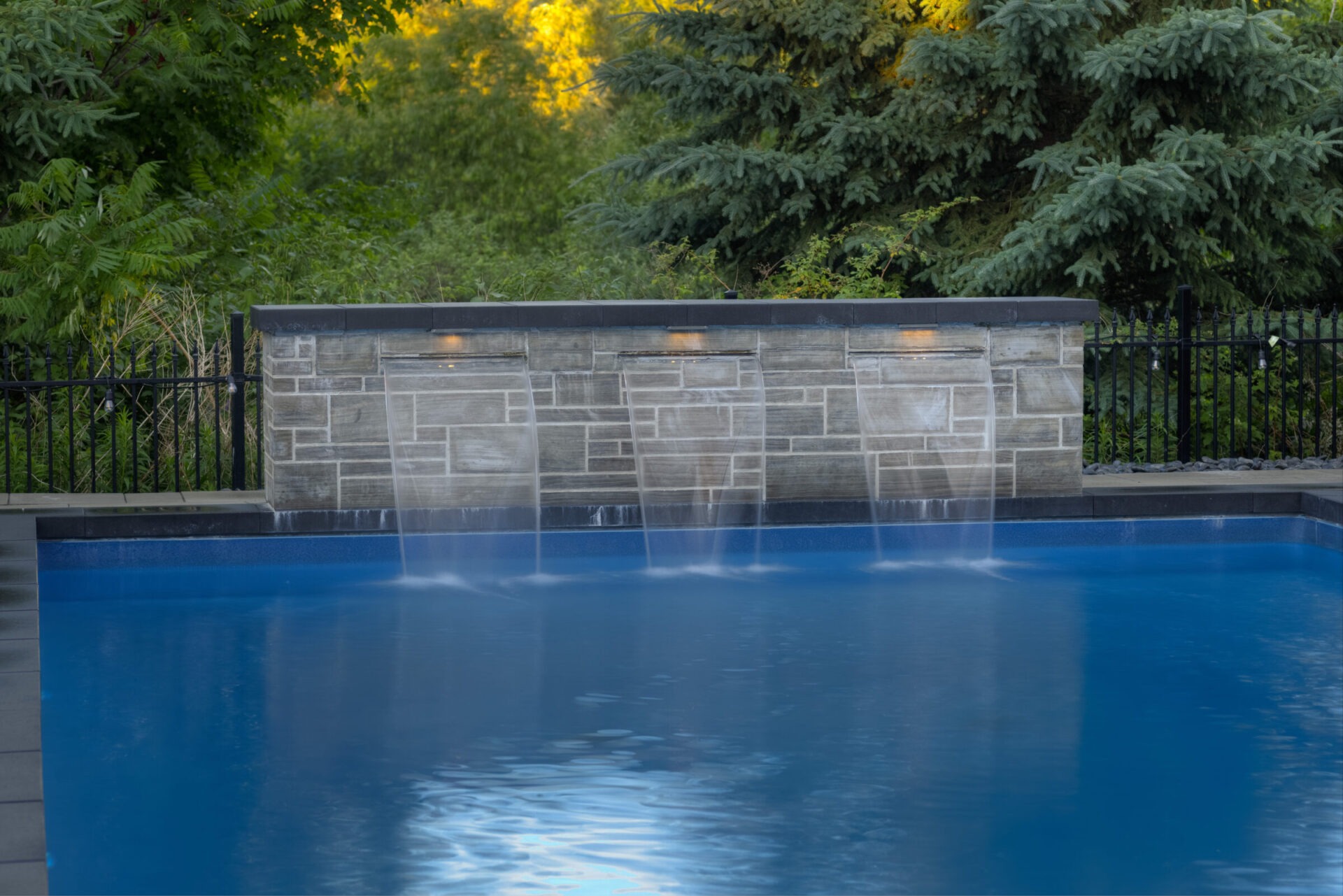 A serene swimming pool with water features cascading from a stone wall, surrounded by lush greenery and fencing, in a tranquil garden setting.