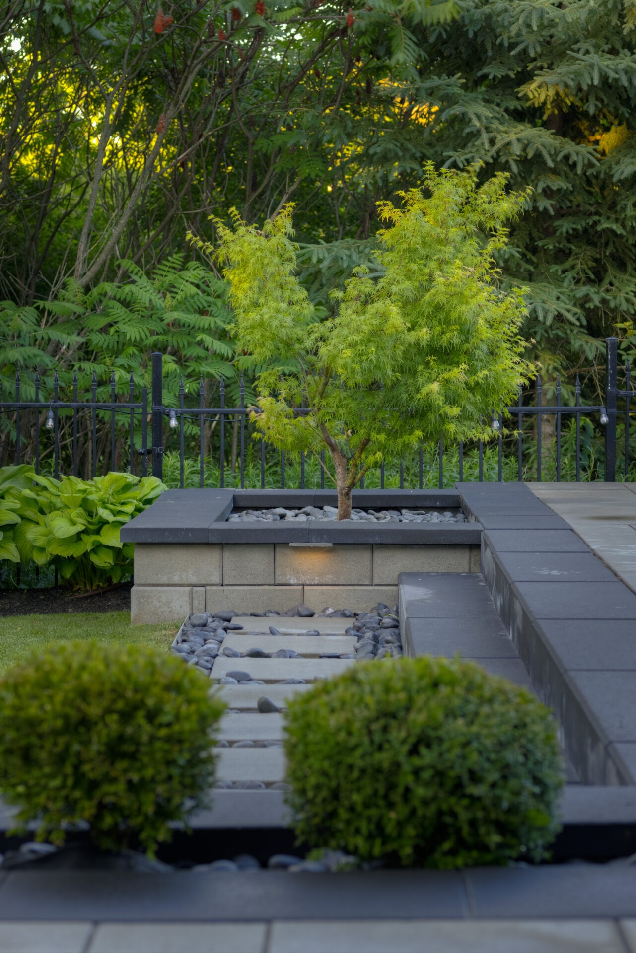 A serene garden pathway with symmetrically placed shrubs leading to a small tree surrounded by stones, accented by understated outdoor lighting, and a lush backdrop.