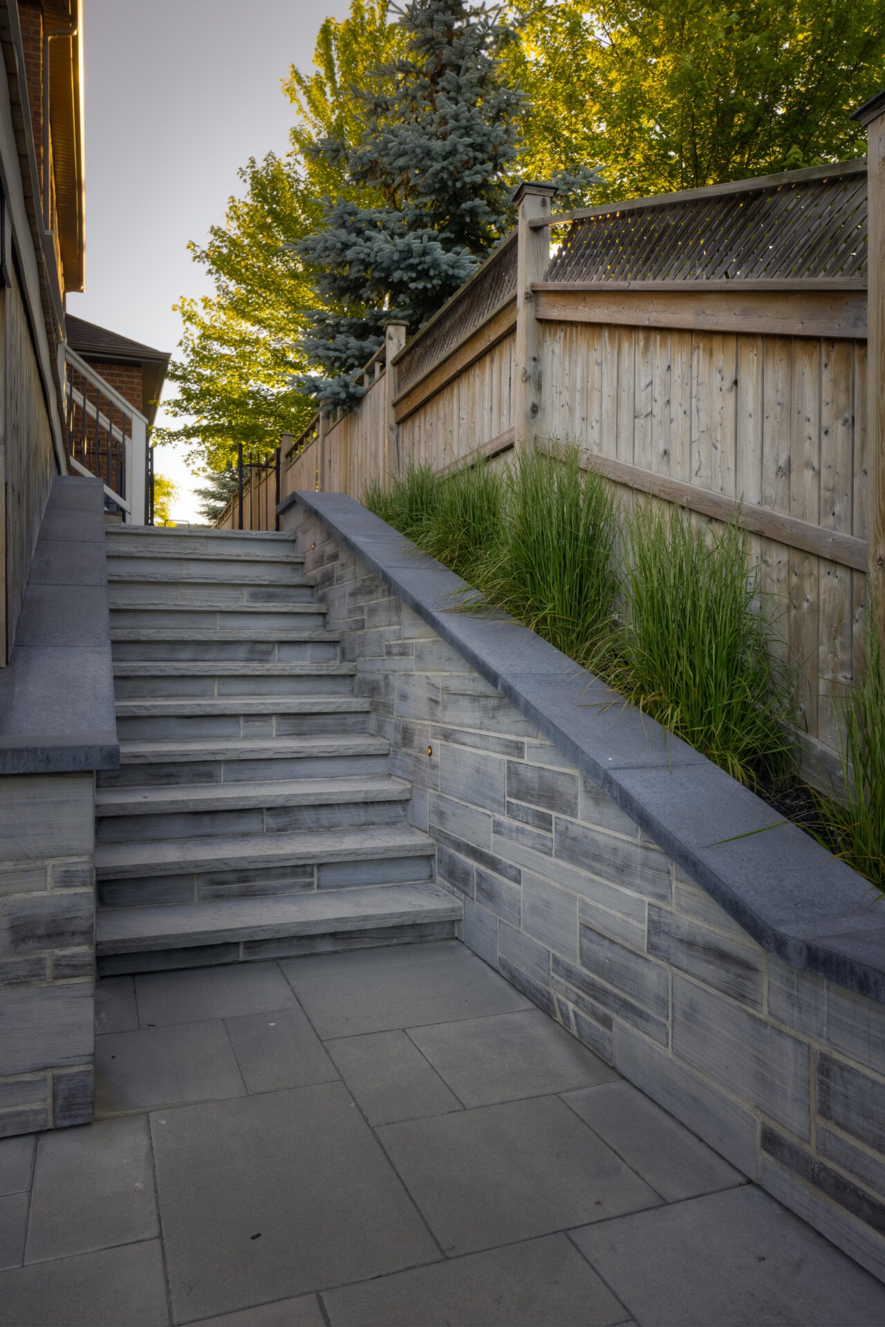 This image shows a set of outdoor steps made of gray stone tiles, framed by a wooden fence and accented with green ornamental grasses.