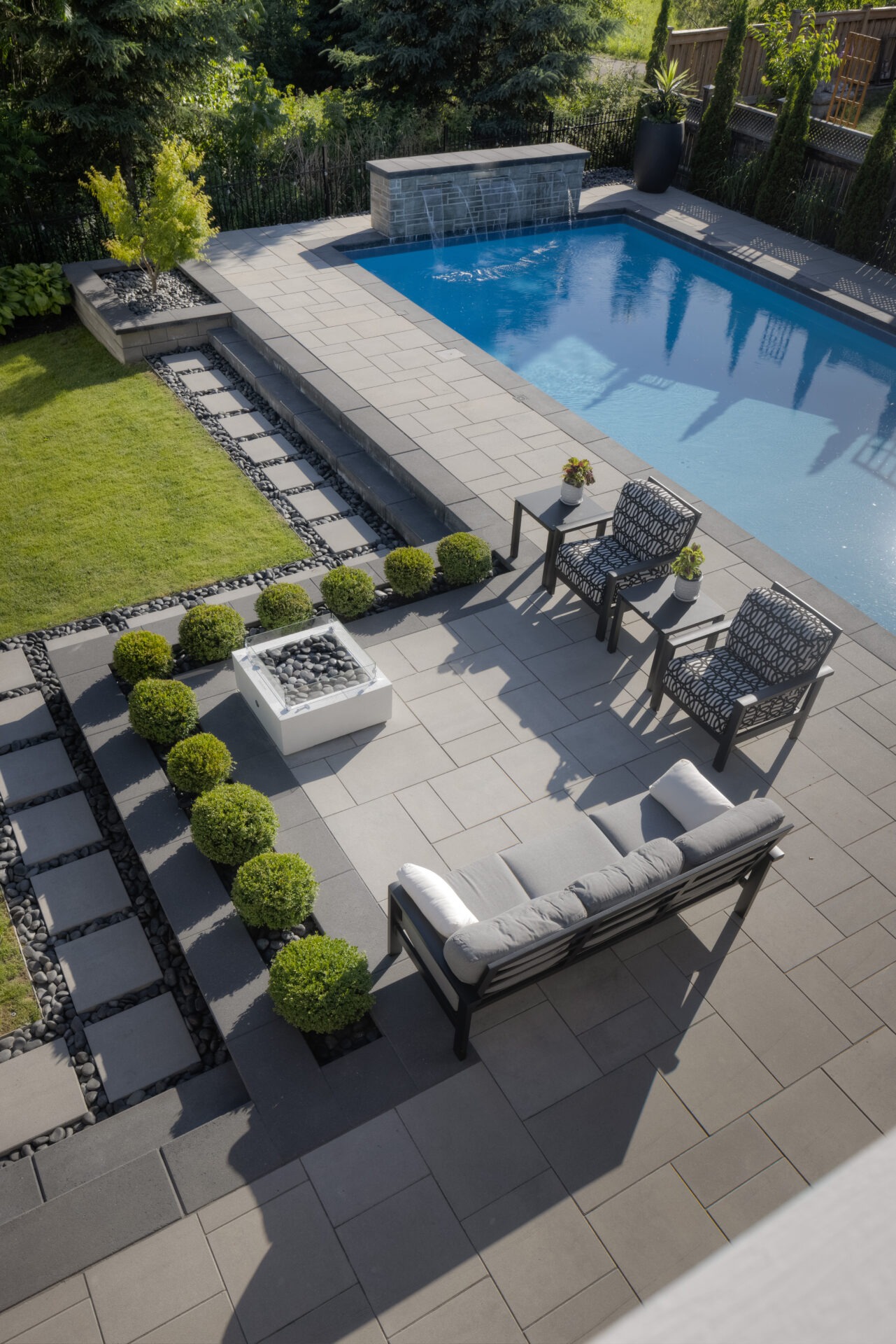 An outdoor area featuring a swimming pool, patio with lounging furniture, neatly arranged shrubbery, a water feature, and a well-maintained lawn.