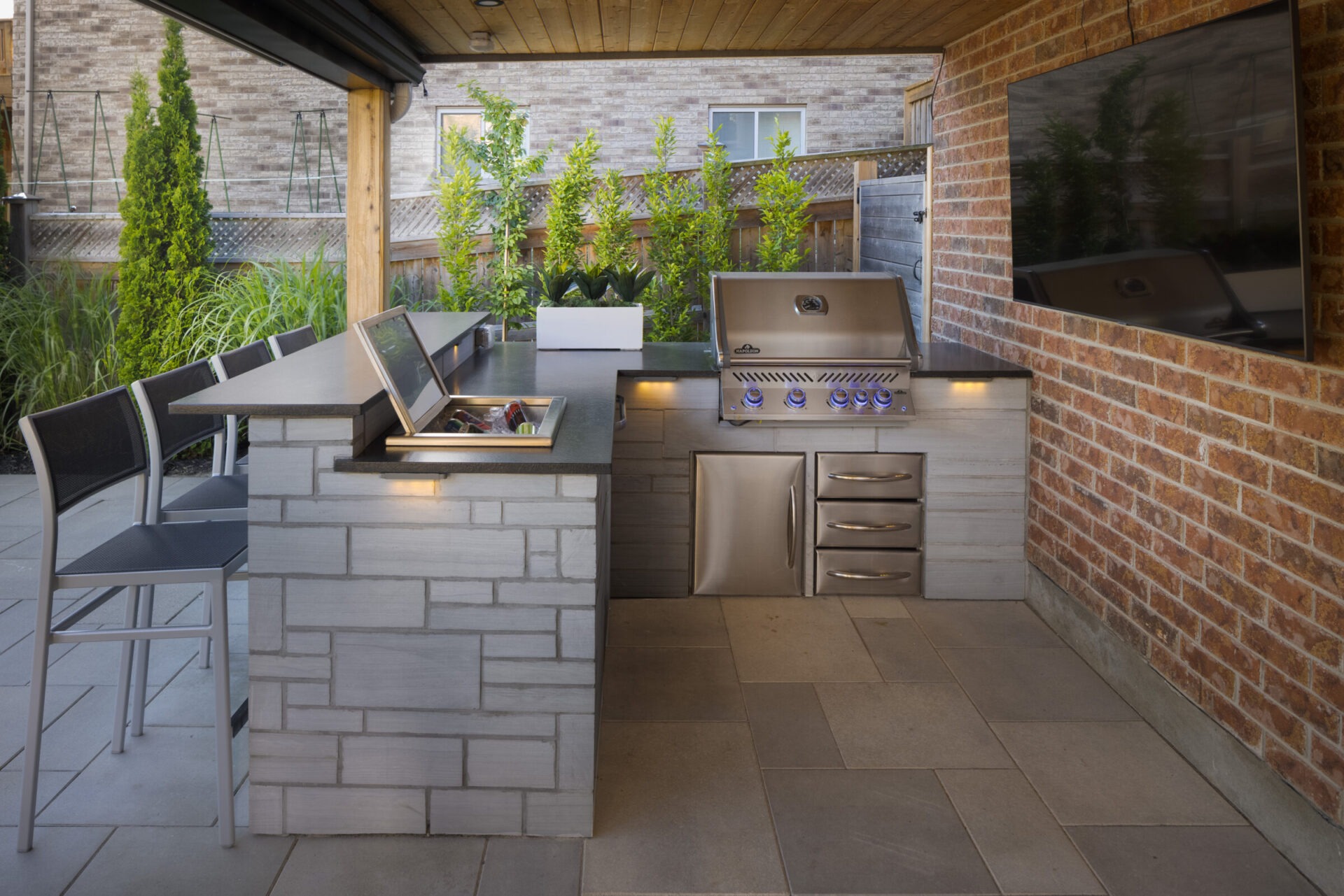 An outdoor kitchen with modern appliances, including a grill and refrigerator, complemented by a bar seating area, under a covered patio with greenery.