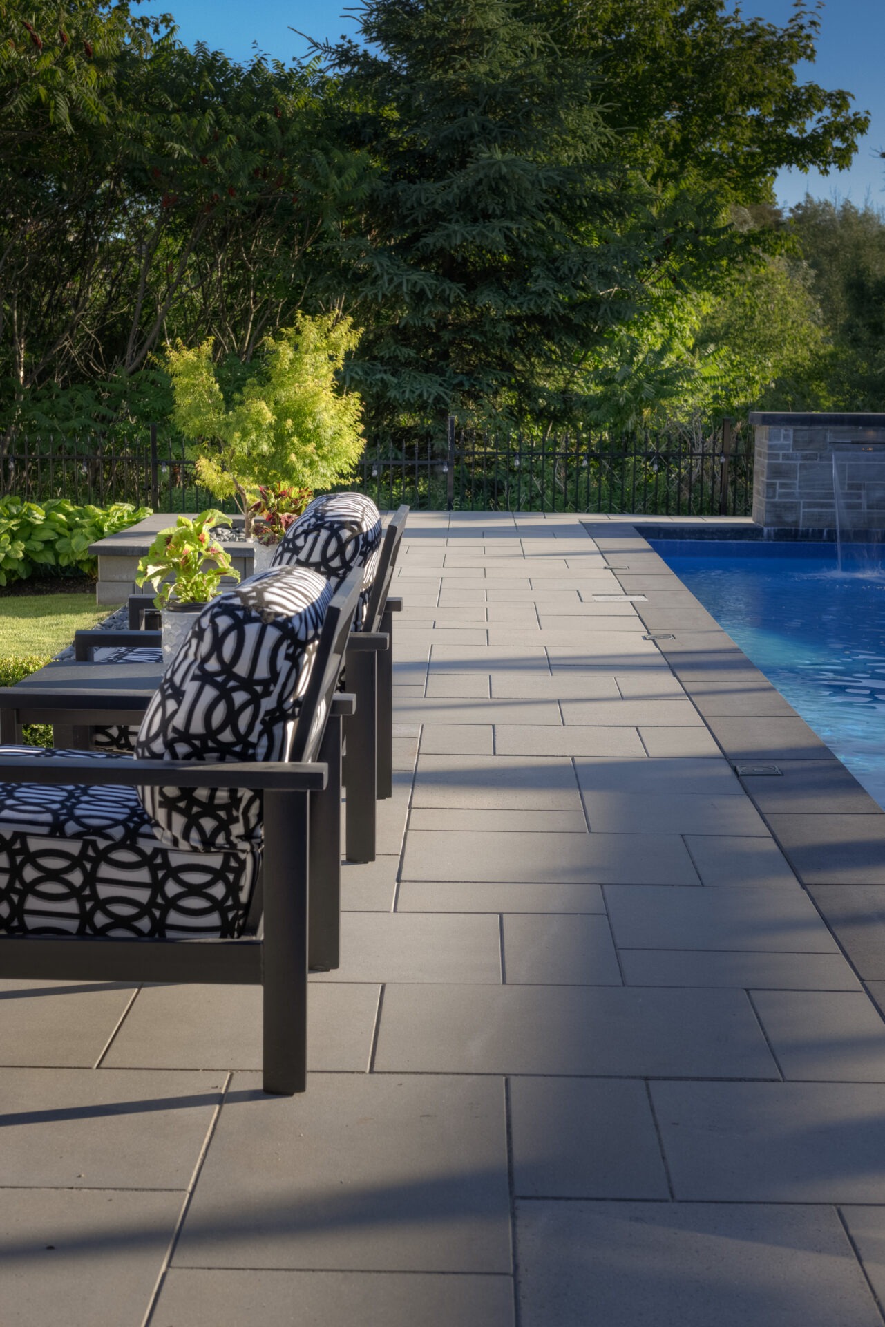 An outdoor poolside area featuring a row of loungers with patterned cushions, a neatly tiled deck, lush greenery, clear skies, and a tranquil setting.