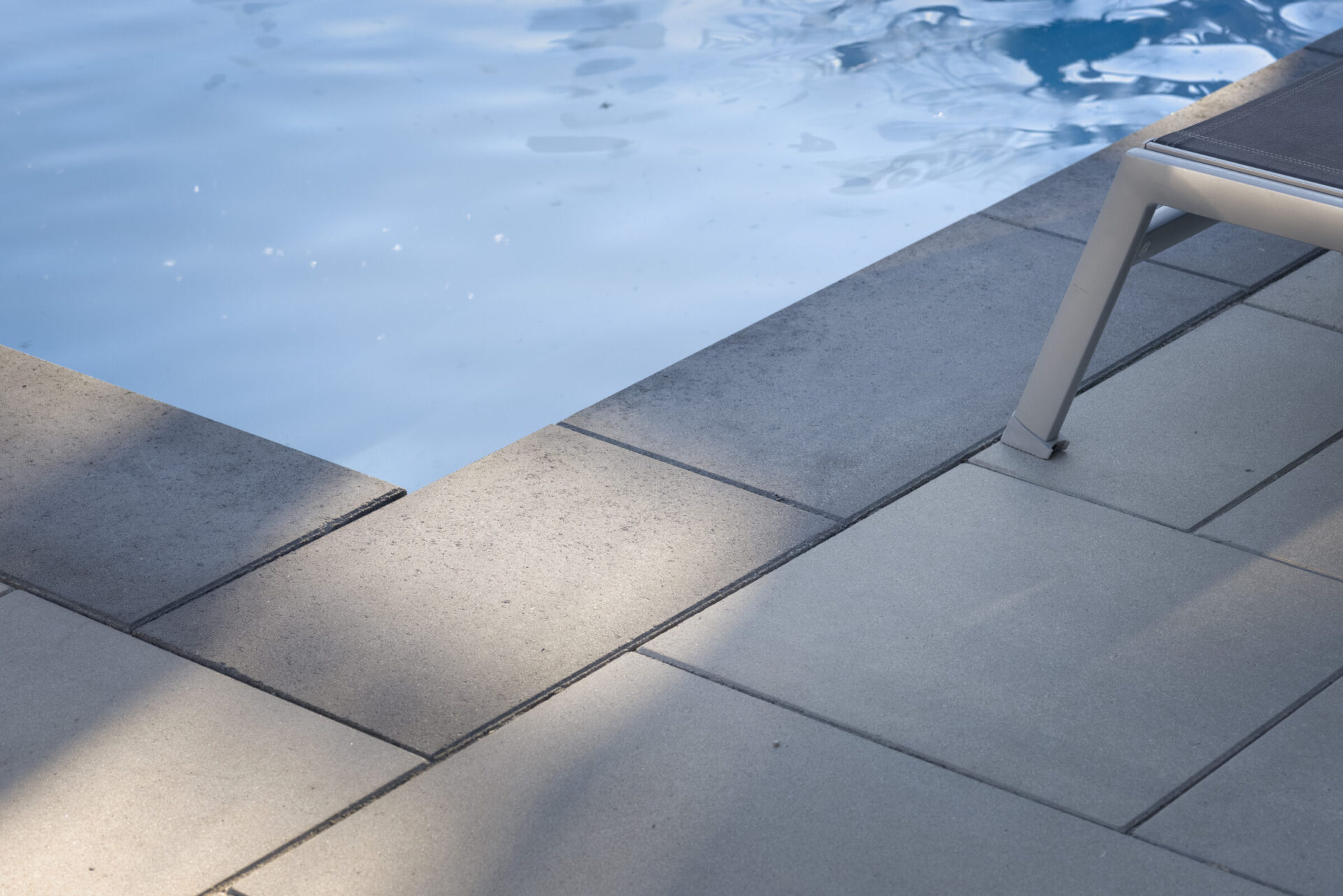 A tranquil scene showing a swimming pool edge with part of a sun lounger visible, clear water with gentle ripples, and a tiled deck.