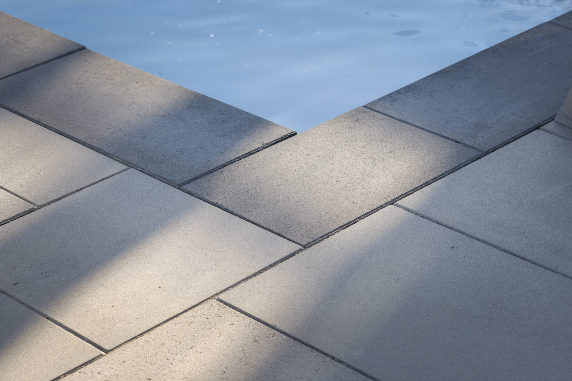 This image shows an edge where tiled flooring meets the calm water of a pool, casting shadows and reflecting the sky above.