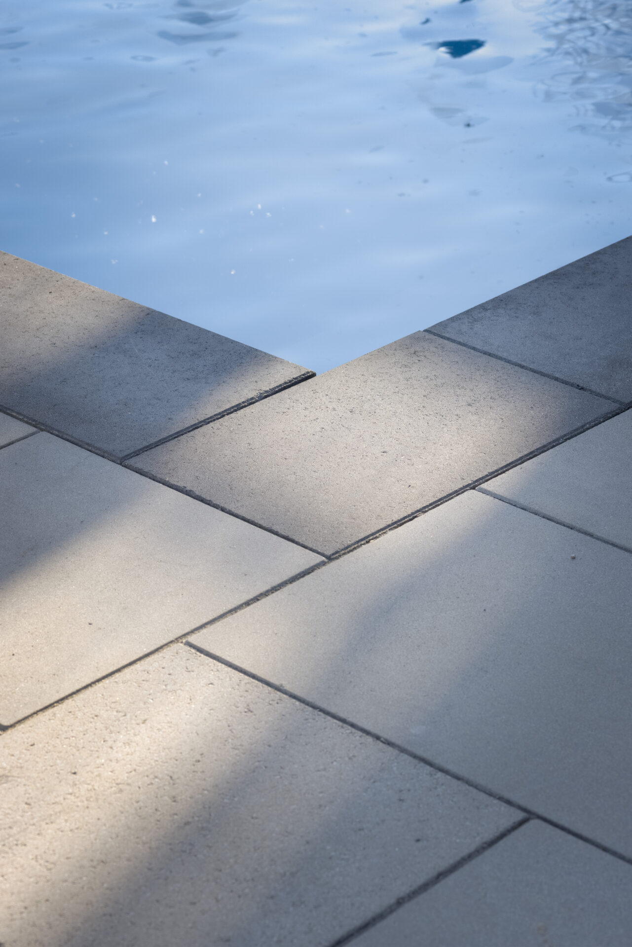 This image shows a pool edge with geometric paving stones leading up to tranquil blue water reflecting sunlight, creating a serene outdoor scene.