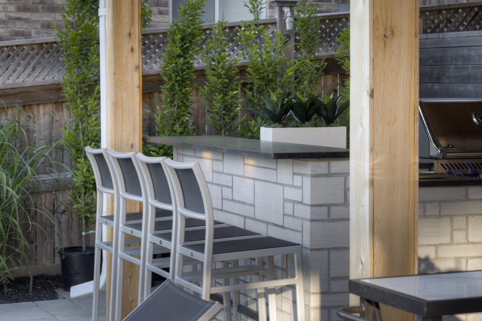 An outdoor patio area with modern bar stools, a built-in grill, wooden accents, greenery, and a privacy fence in a residential backyard setting.