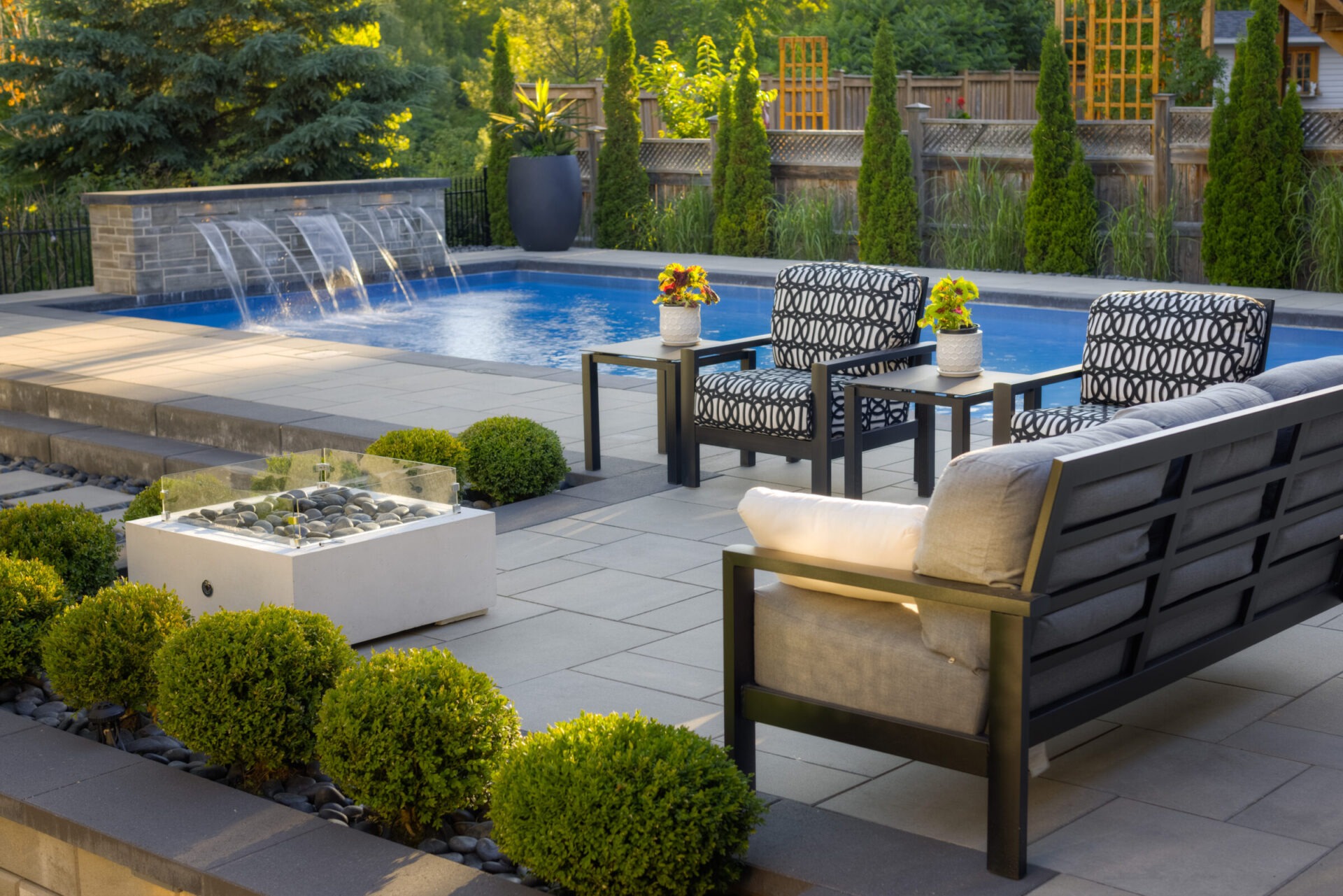 An inviting backyard with a rectangular blue swimming pool, water features, stylish outdoor furniture, manicured bushes, and a stone patio.