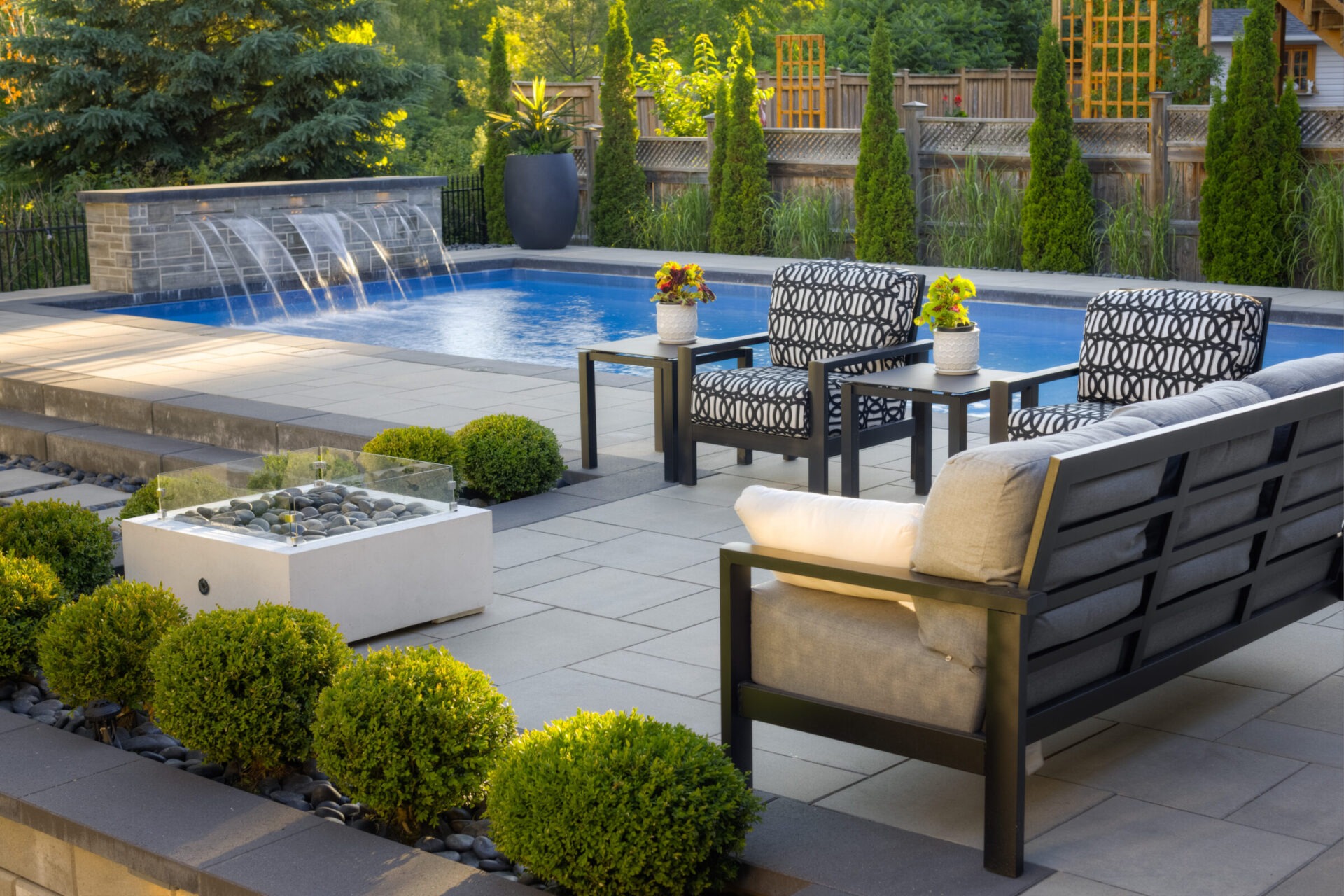 An elegant outdoor patio with a swimming pool featuring waterfalls, surrounded by lush plants, modern furniture, and a fire feature for ambiance.