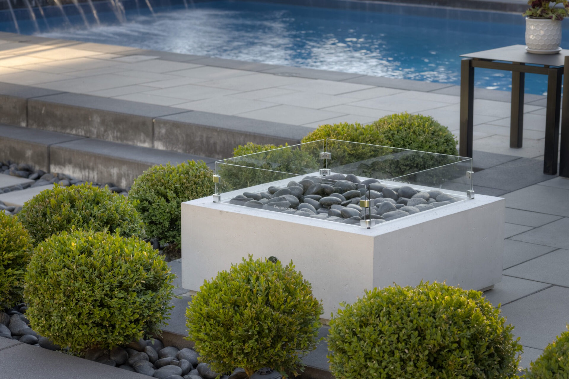 This image features a modern outdoor setting with a fire pit, rounded stones, boxwood shrubs, a swimming pool with fountains, and a side table.