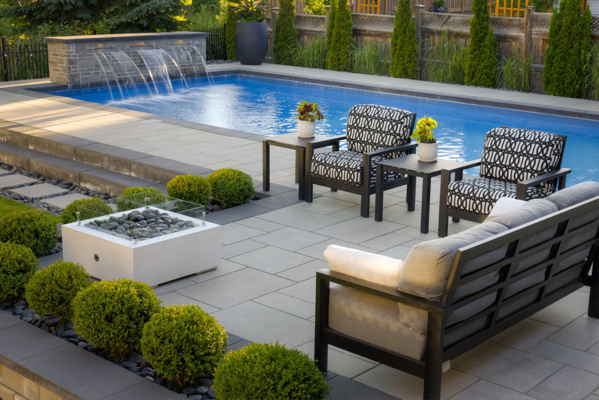 An elegant outdoor patio features patterned chairs, a modern fireplace, neatly trimmed shrubs, a swimming pool with fountains, and a tiled floor.