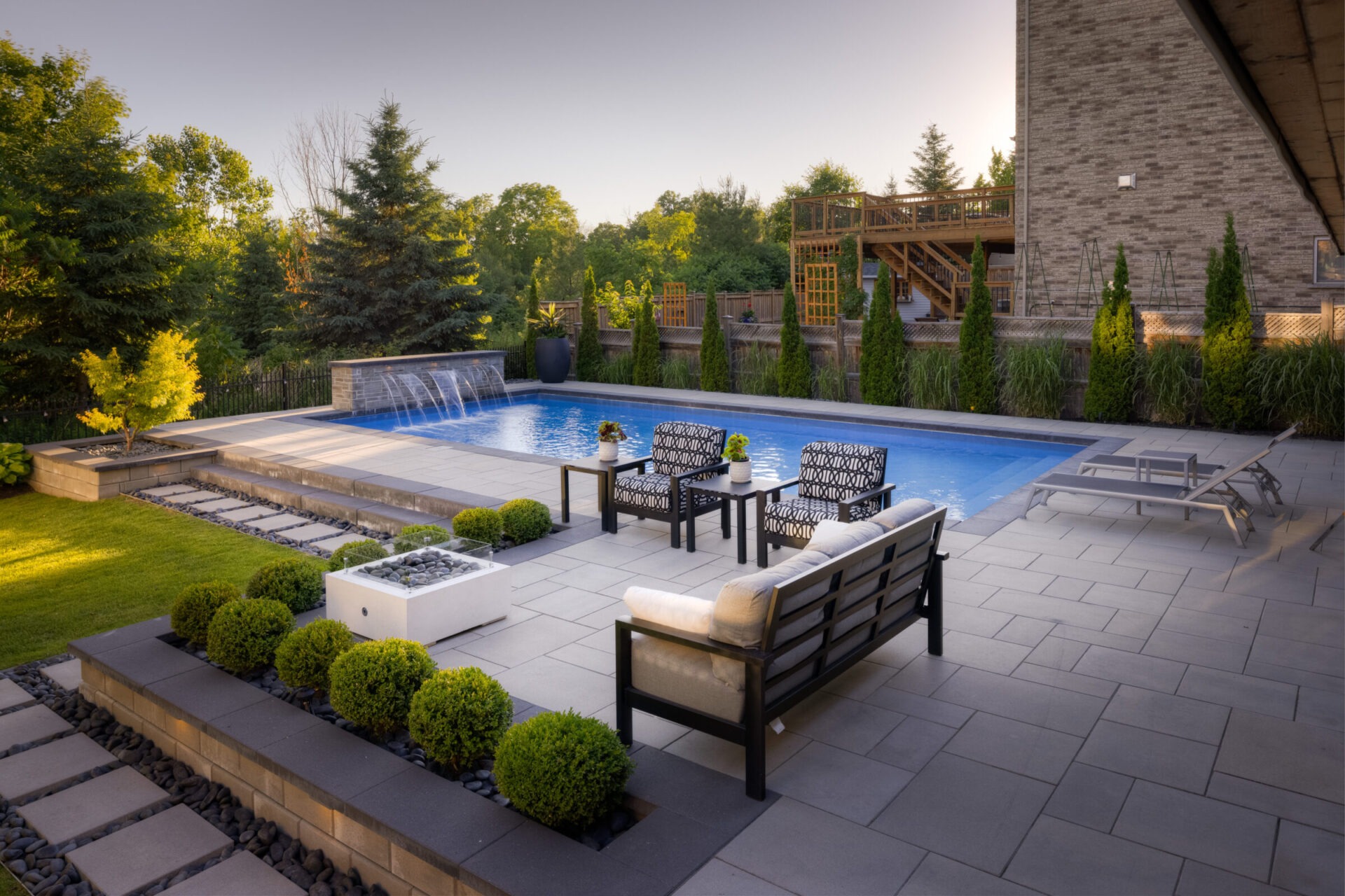 An elegant backyard featuring a swimming pool with fountains, a patio with comfortable seating, manicured greenery, and a raised wooden deck in a tranquil setting.
