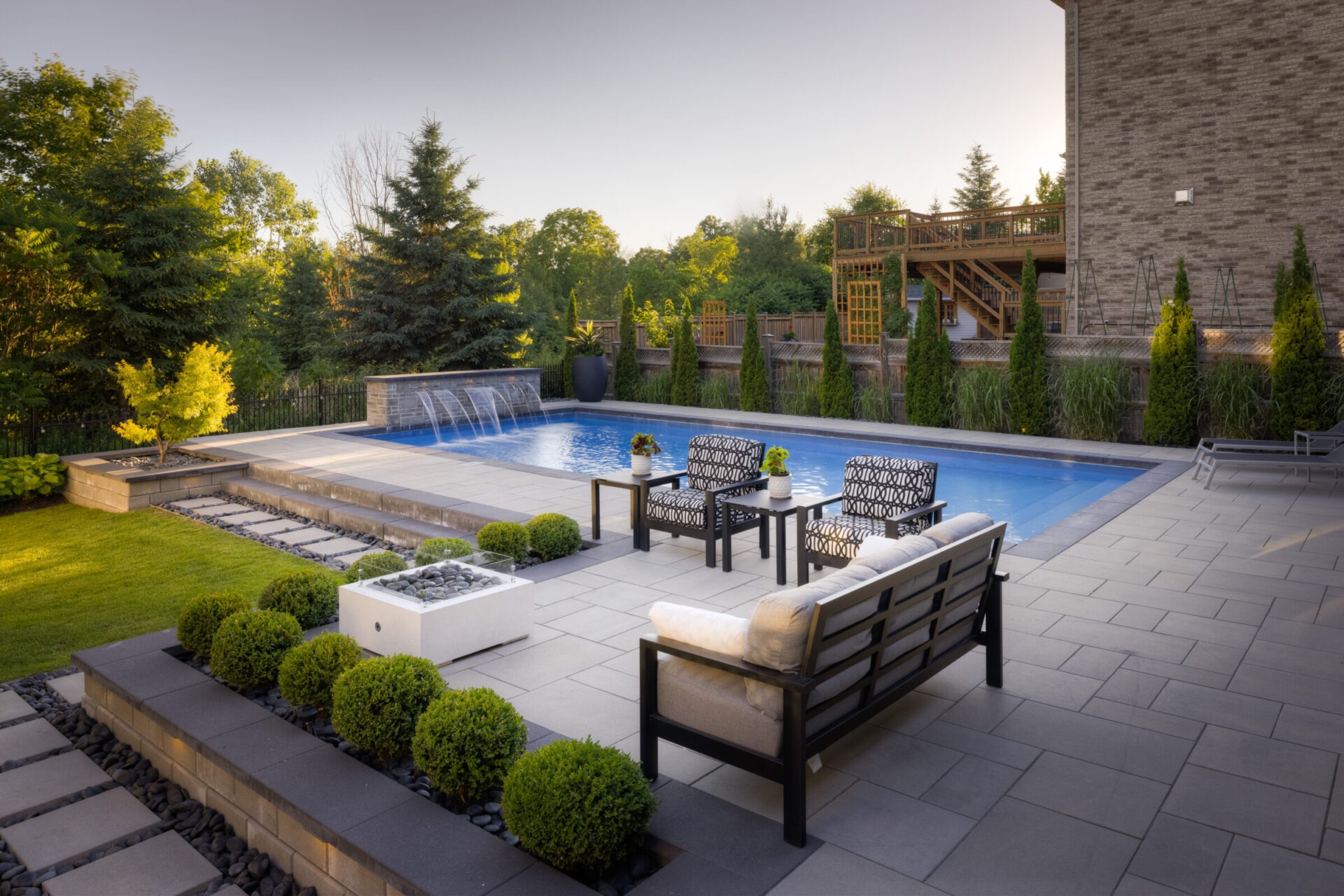 A well-landscaped backyard with a swimming pool, water feature, patio furniture, neatly trimmed bushes, and a wooden deck amid lush greenery and clear skies.