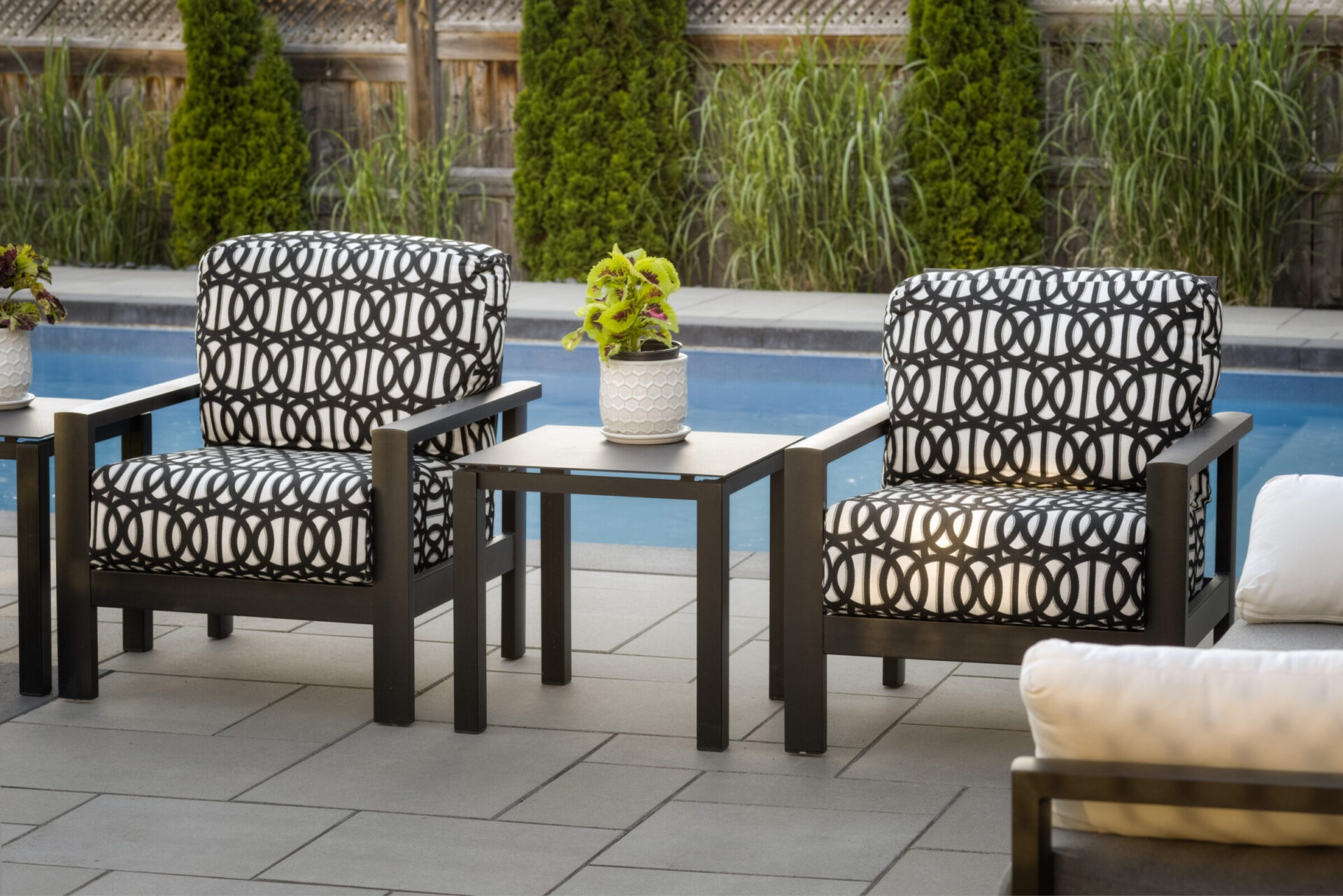 Two modern armchairs with geometric patterned cushions on a patio beside a swimming pool. A small table with a potted plant sits between them.