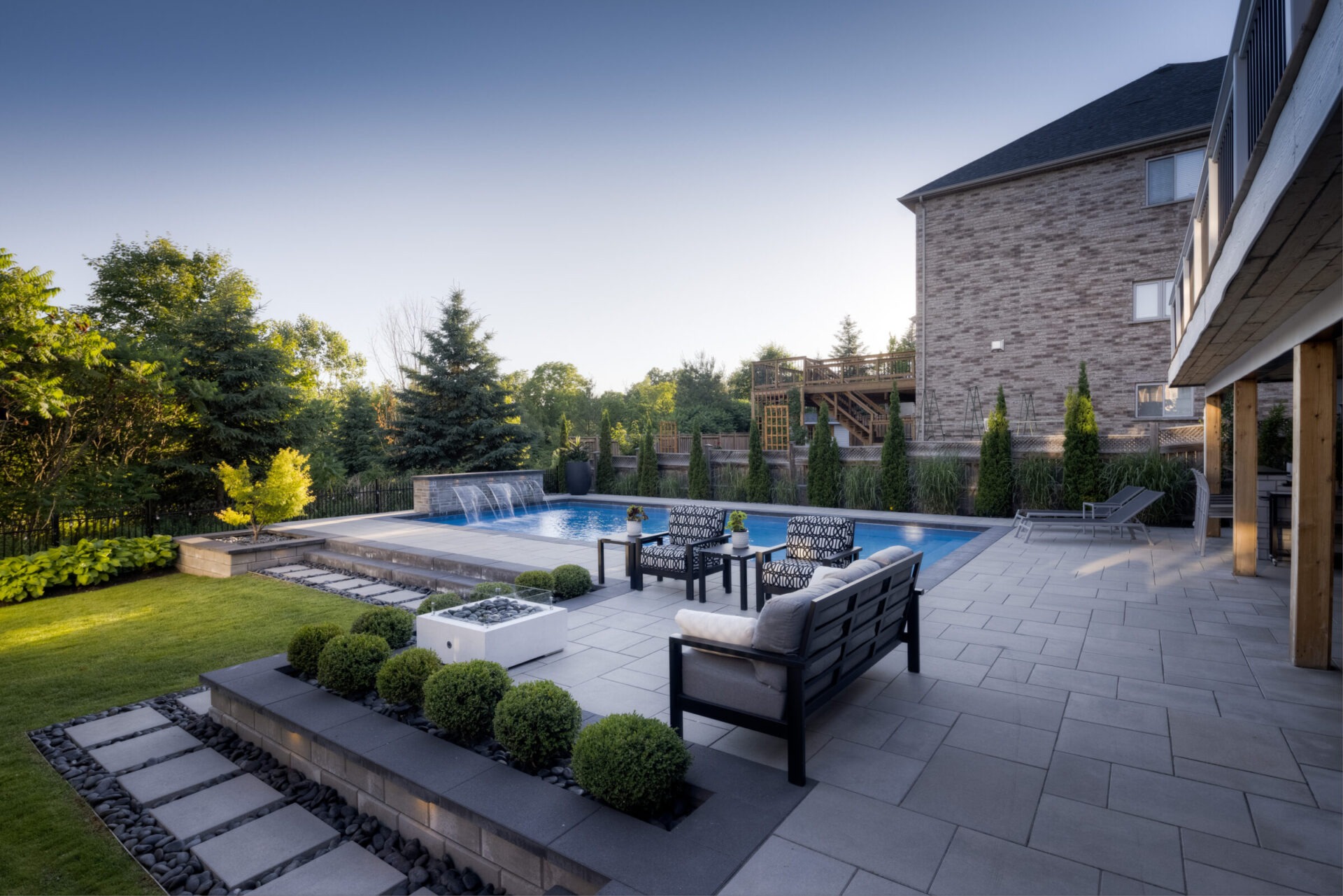 An elegant backyard featuring a swimming pool with a fountain, modern patio furniture, landscaped garden, and a wooden deck under a clear blue sky.