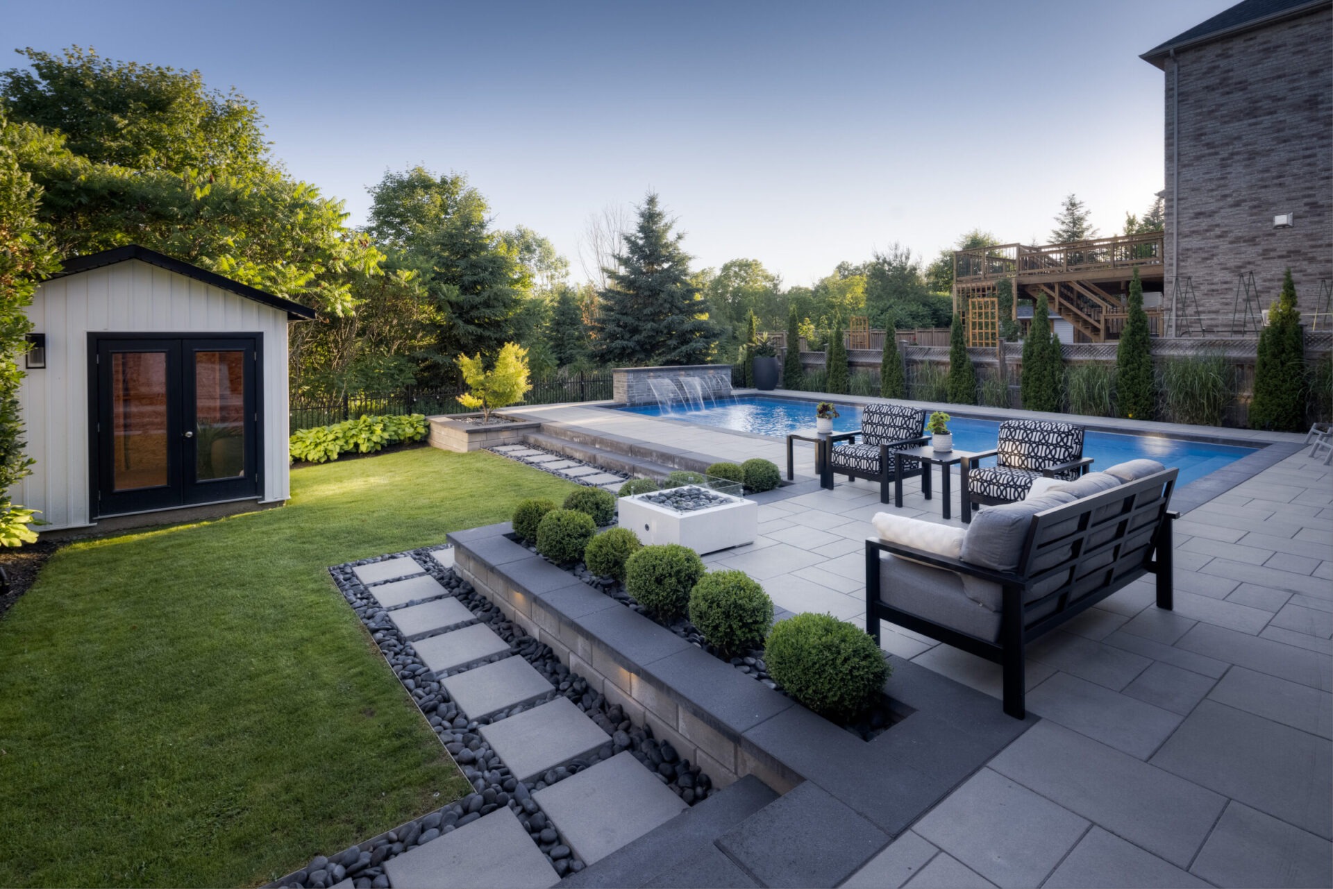 This is a modern backyard with a swimming pool, outdoor seating, manicured lawn, and landscaping. A small shed and a wooden deck are also visible.