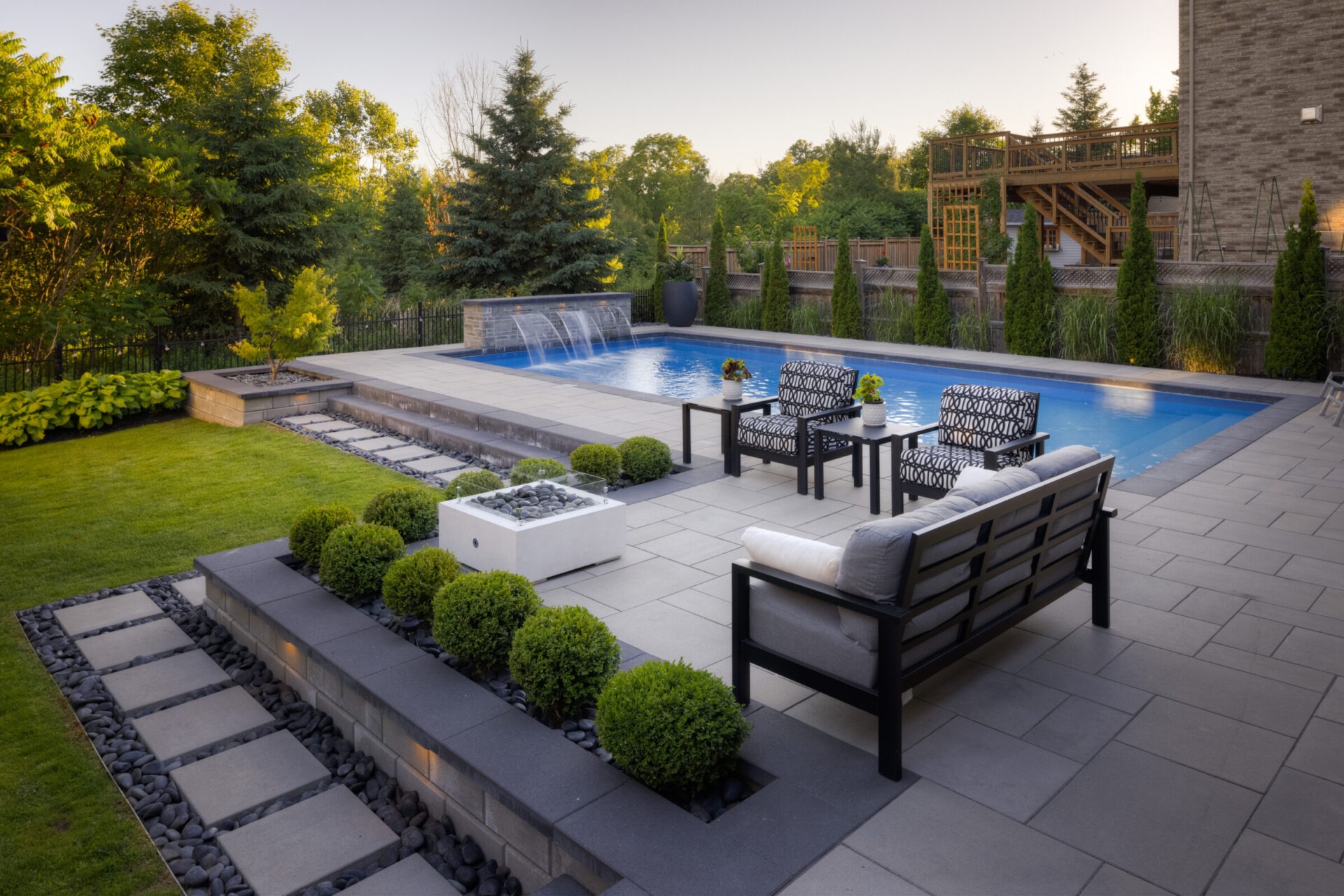 A luxurious backyard with a patio area featuring modern outdoor furniture, an in-ground pool, manicured greenery, and a tranquil water feature.
