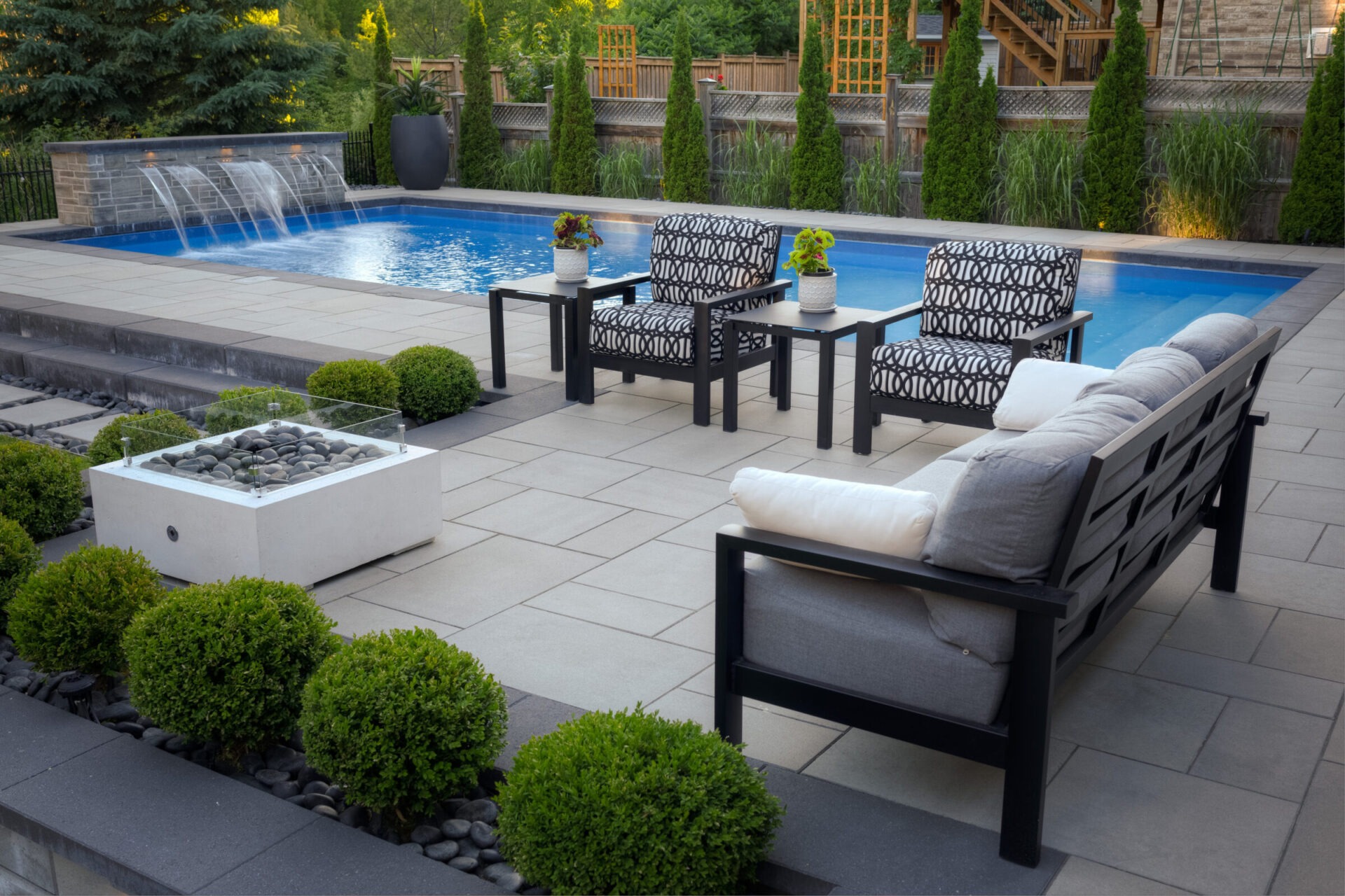 This image shows a luxurious outdoor patio with a blue pool, waterfall feature, stylish seating, landscaped bushes, and a fire pit table.
