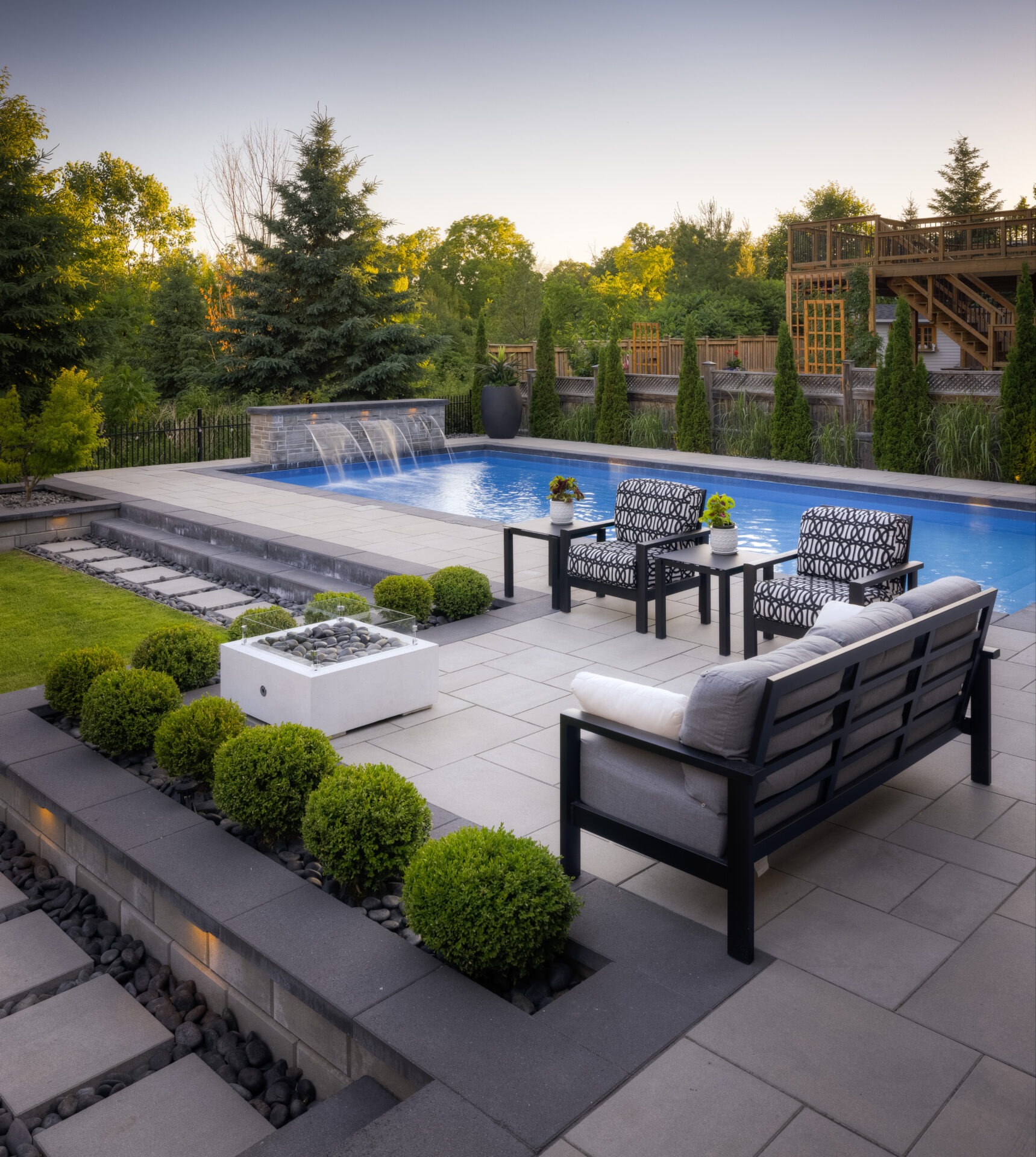 An elegant outdoor area with a swimming pool, water feature, modern patio furniture, landscaped garden, and a wooden deck in a serene setting.