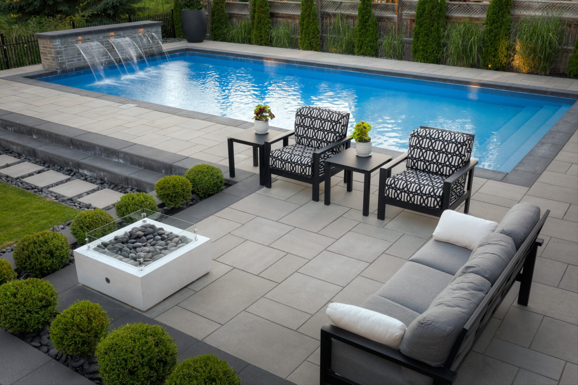 This image shows a modern backyard patio with well-manicured grass, a rectangular inground pool, stylish outdoor furniture, decorative plants, and a water feature.