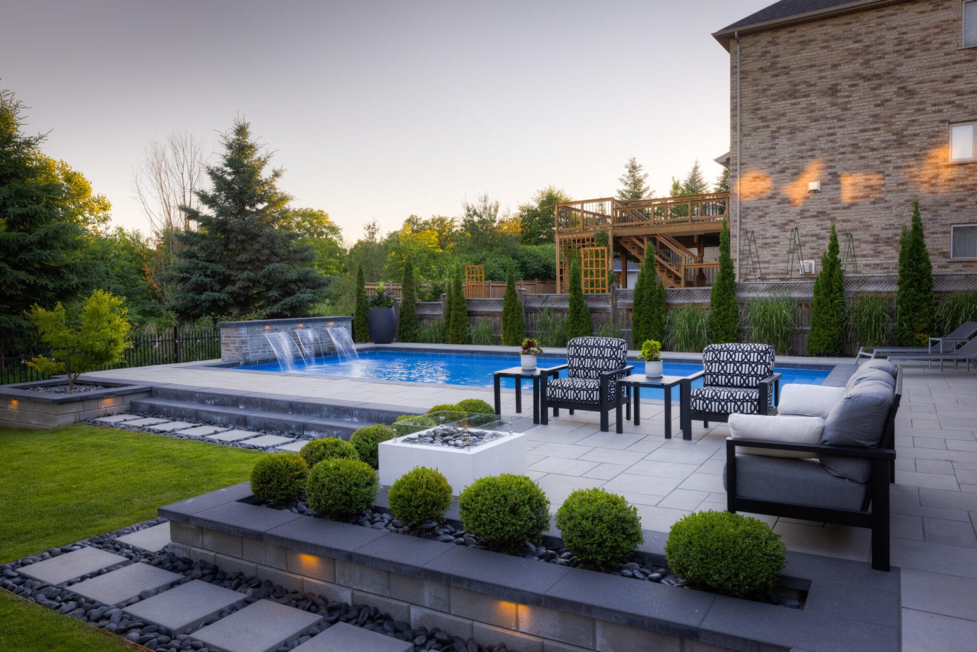 This image shows an elegant backyard with a swimming pool, waterfall feature, stylish outdoor furniture, manicured bushes, and a wooden deck.