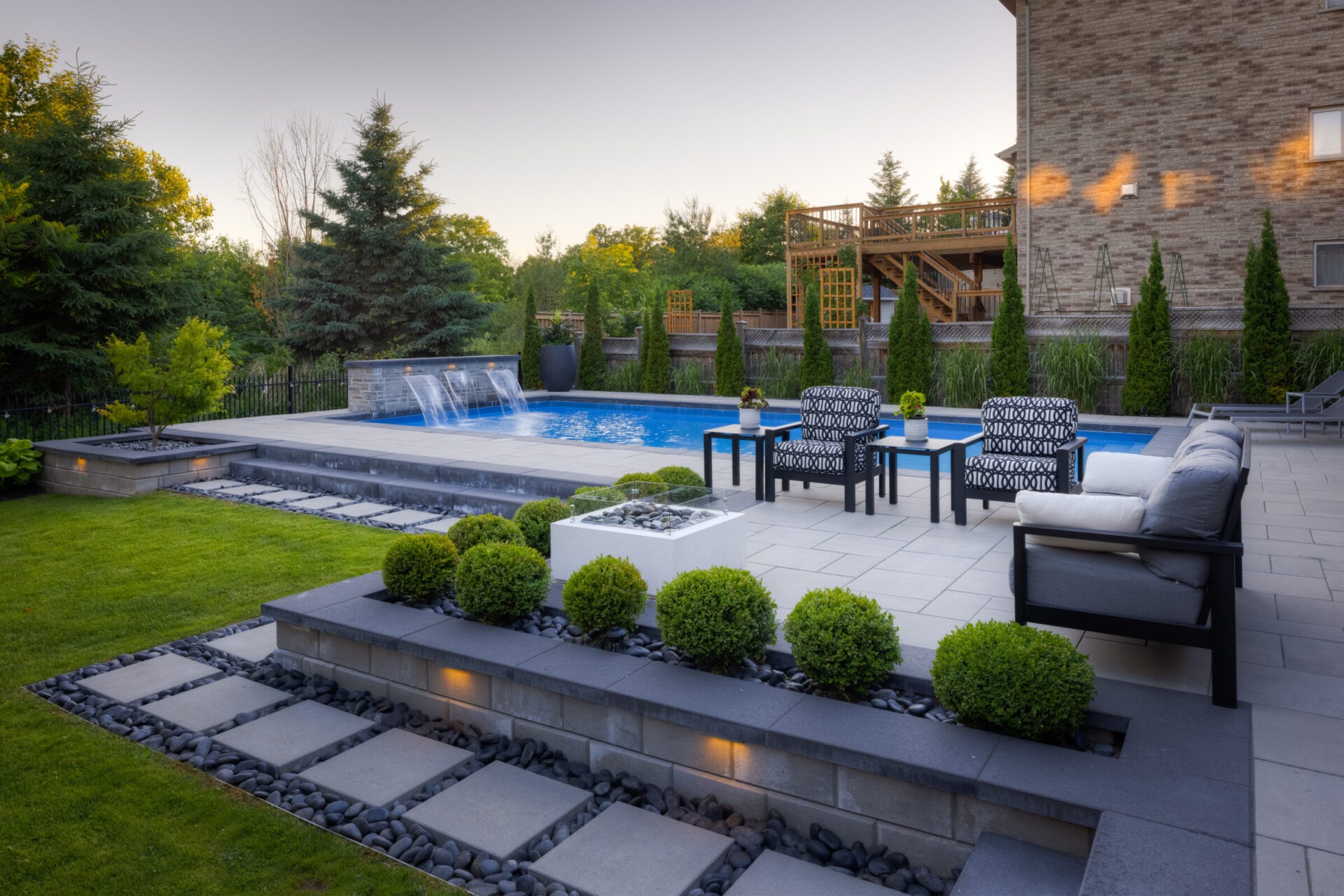 An elegant backyard with a swimming pool, waterfall, patio furniture, manicured shrubs, and stepping stones, surrounded by trees and a wooden deck.