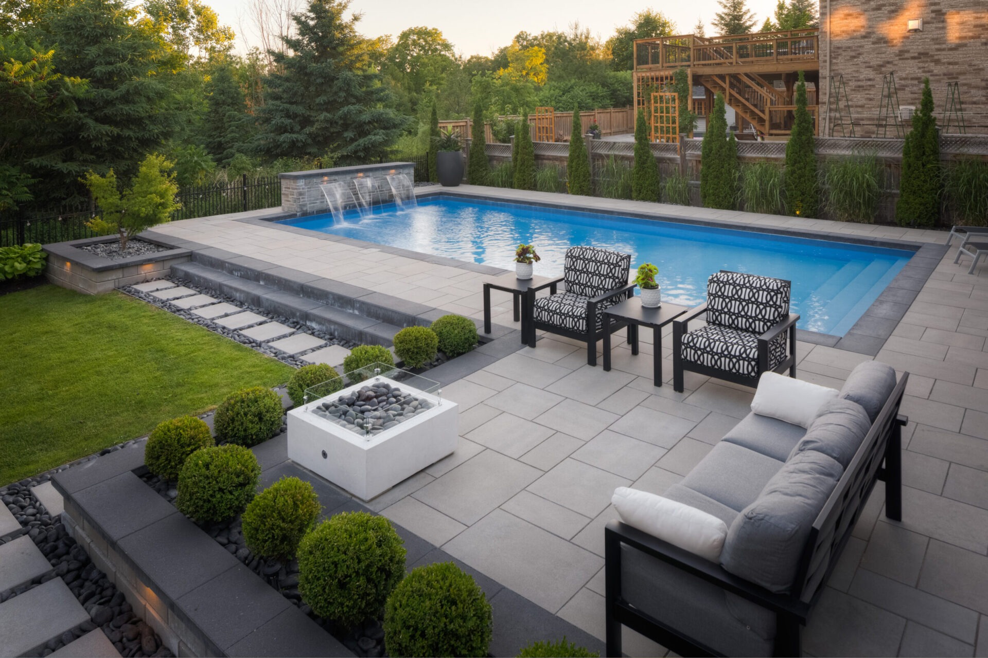 An elegant backyard with a large swimming pool, stylish patio furniture, a fire pit, meticulously landscaped greenery, and a wooden deck under twilight sky.