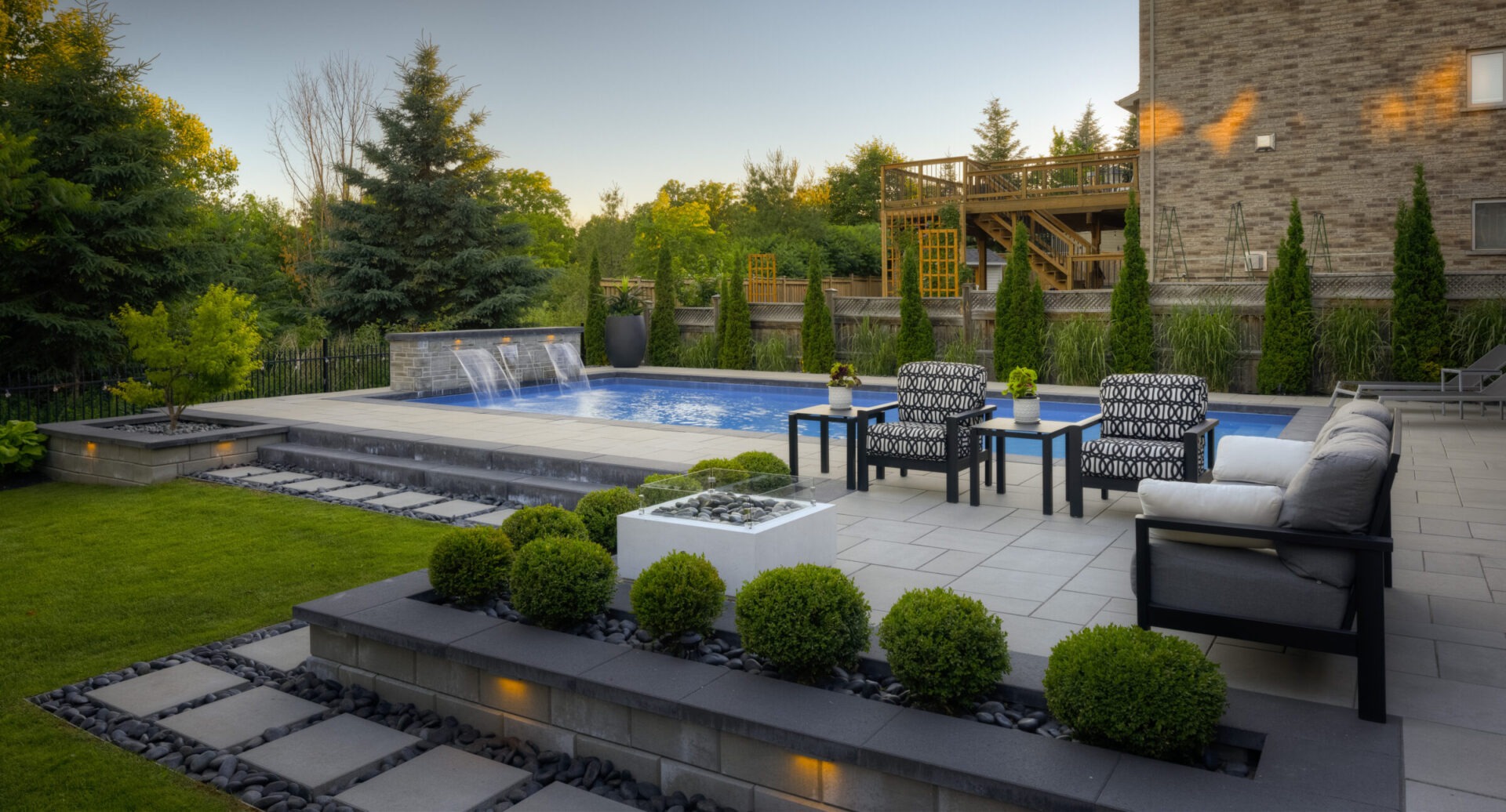 An elegant backyard with a swimming pool, fountain, modern furniture, manicured bushes, and a two-level wooden deck surrounded by lush greenery at dusk.
