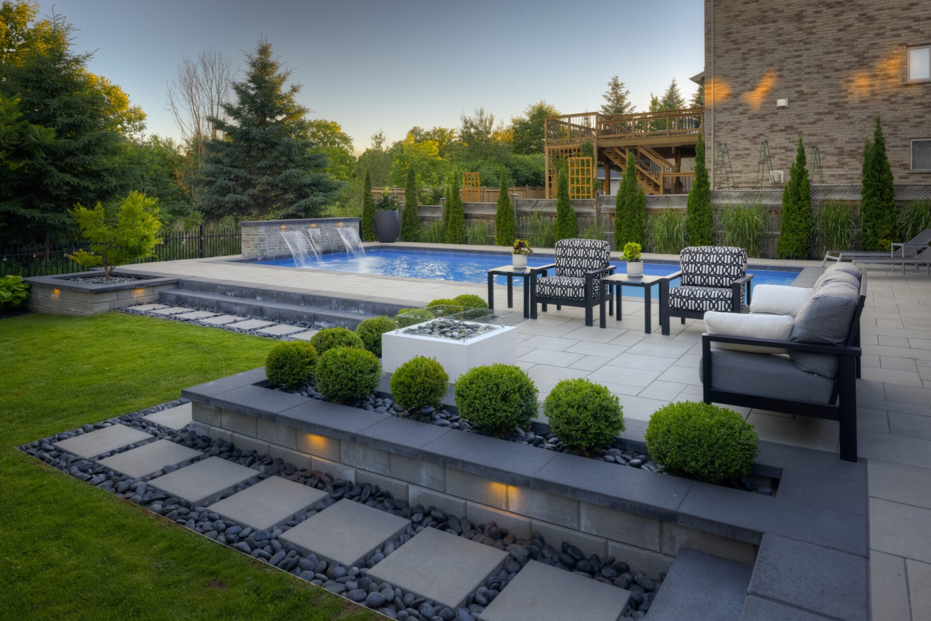An elegantly landscaped backyard with a swimming pool, water features, patio furniture, and a wooden deck surrounded by trees during the late afternoon.