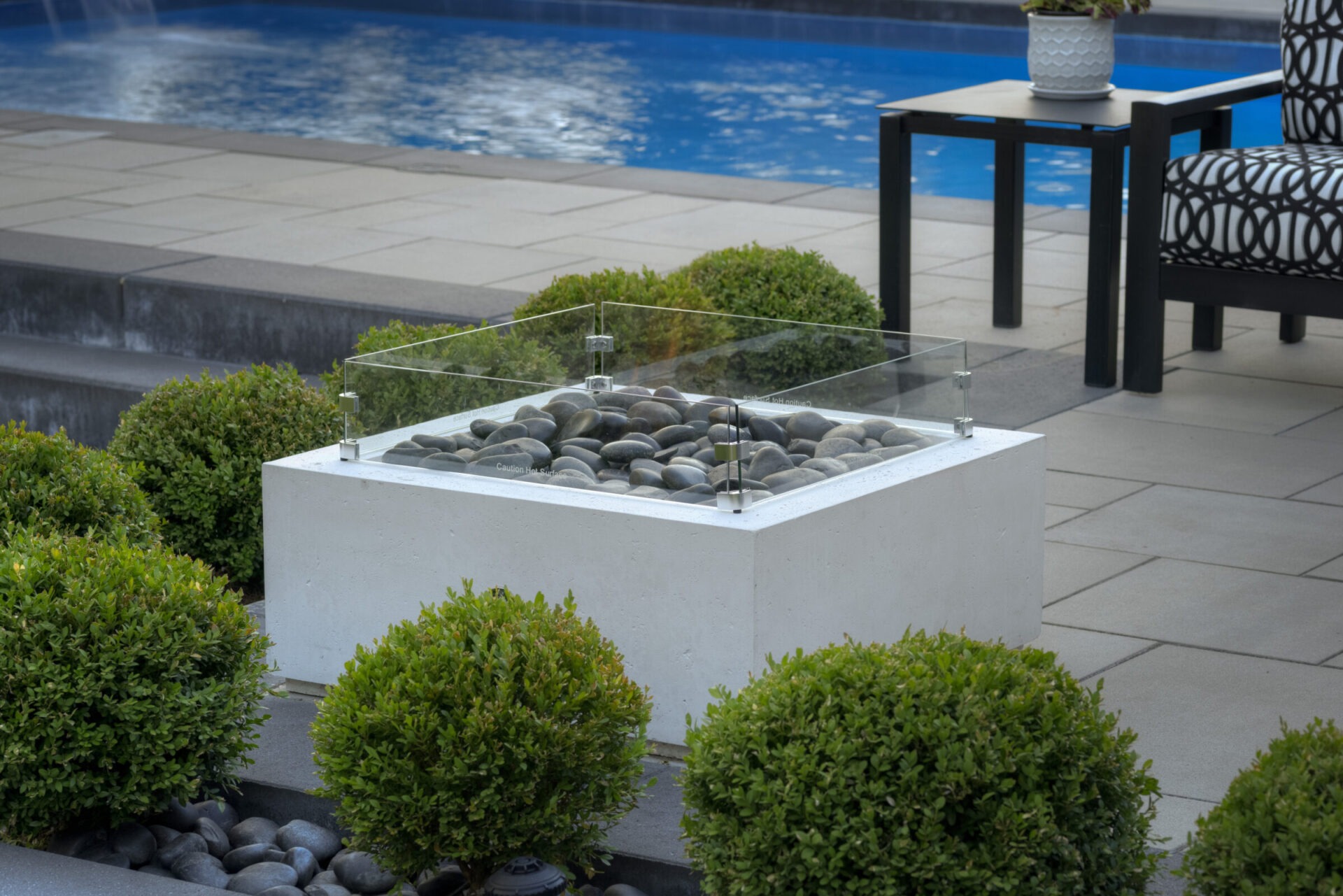 This is an outdoor setting featuring a patio with a fire pit containing grey stones, neat hedges, garden furniture beside a swimming pool.