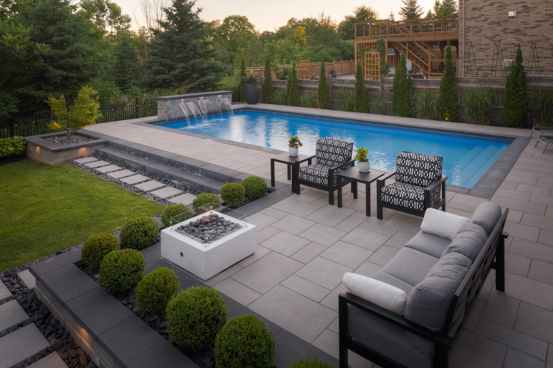 Elegant backyard featuring a blue swimming pool, patio with modern outdoor furniture, landscaped lawn, and a multi-level wooden deck at dusk.