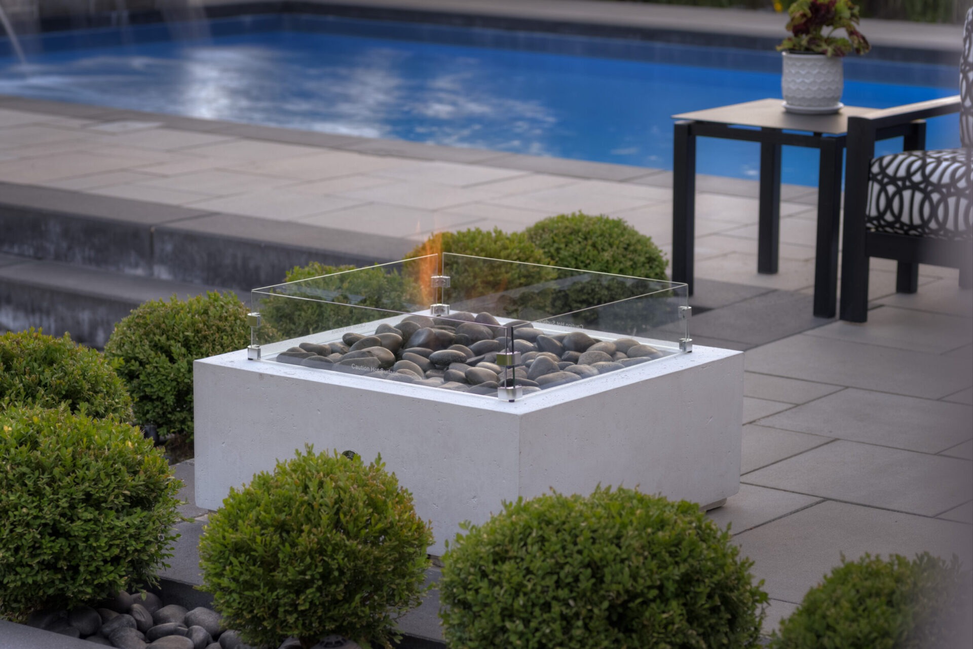 An outdoor fire pit with glass shield sits on a patio, surrounded by shrubs, with a pool and outdoor furniture in the background.
