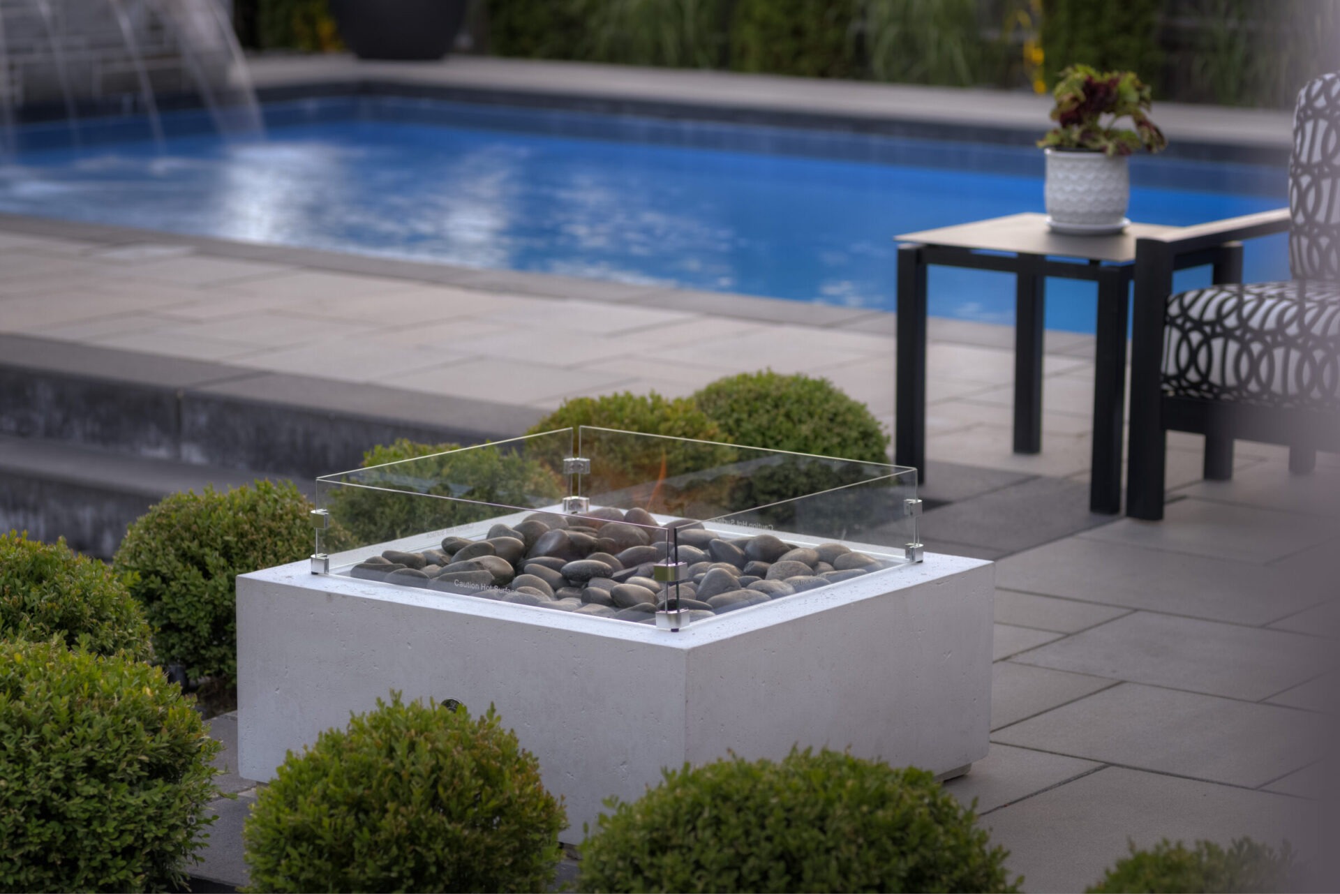 An outdoor setting with a modern fire pit surrounded by rocks, near a pool. Shrubbery, a patio, and outdoor furniture complete the tranquil scene.
