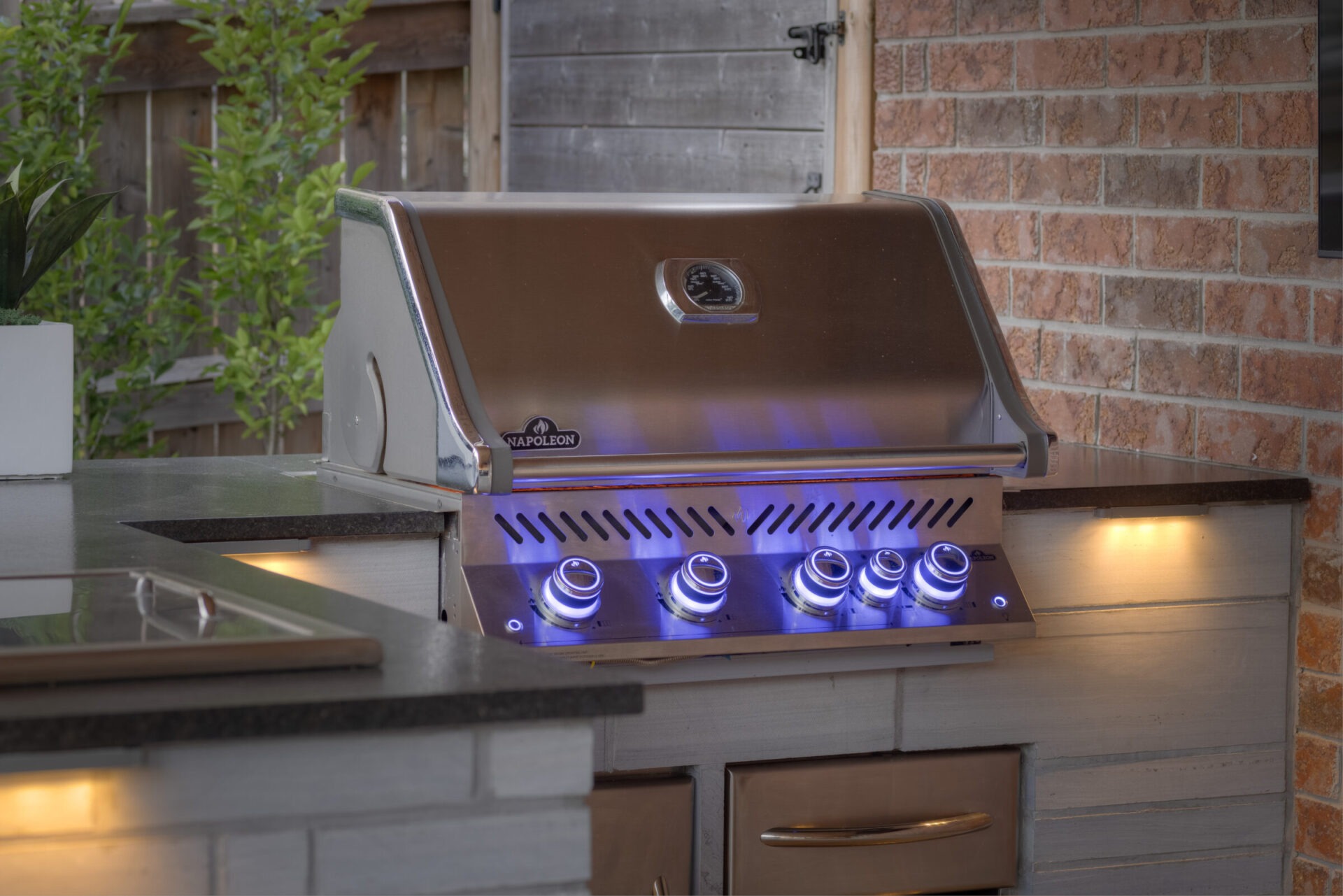 A modern outdoor grill with illuminated control knobs is set against a brick wall in an outdoor kitchen setting surrounded by greenery.