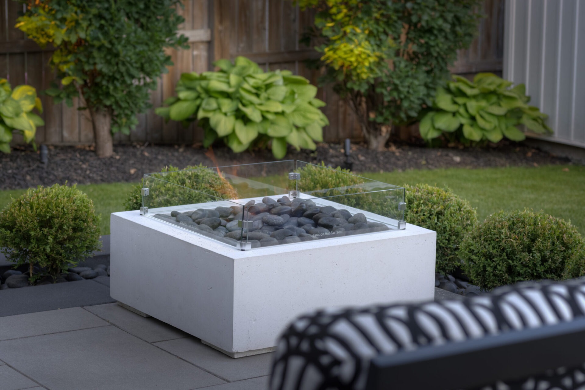A modern outdoor fire pit with glass shield and smooth stones inside, located in a neatly landscaped garden with shrubs, foliage, and a wooden fence.