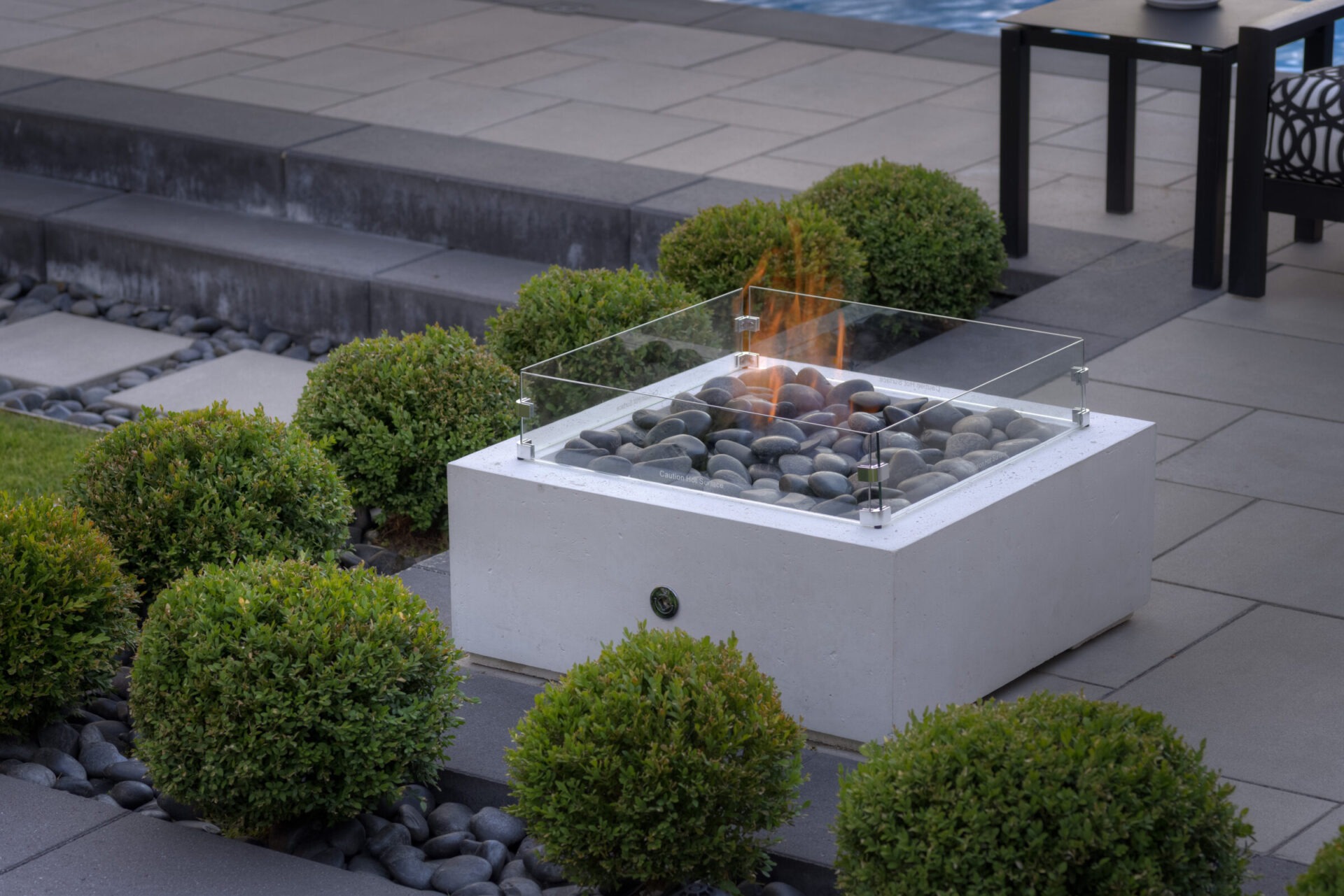 An outdoor gas fire pit with glass wind guard on a patio, surrounded by neatly trimmed green shrubs and smooth grey stones.