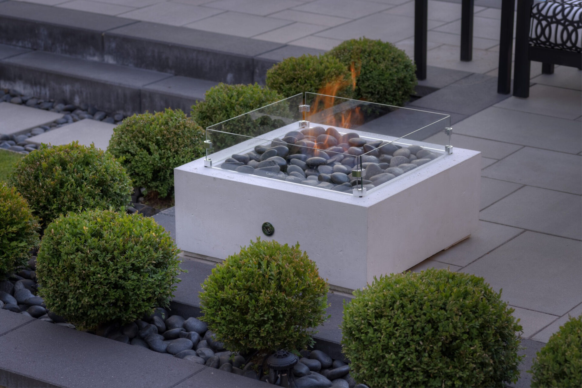 A modern outdoor gas fire pit with a glass shield surrounded by decorative round bushes and smooth gray stones on a patio.