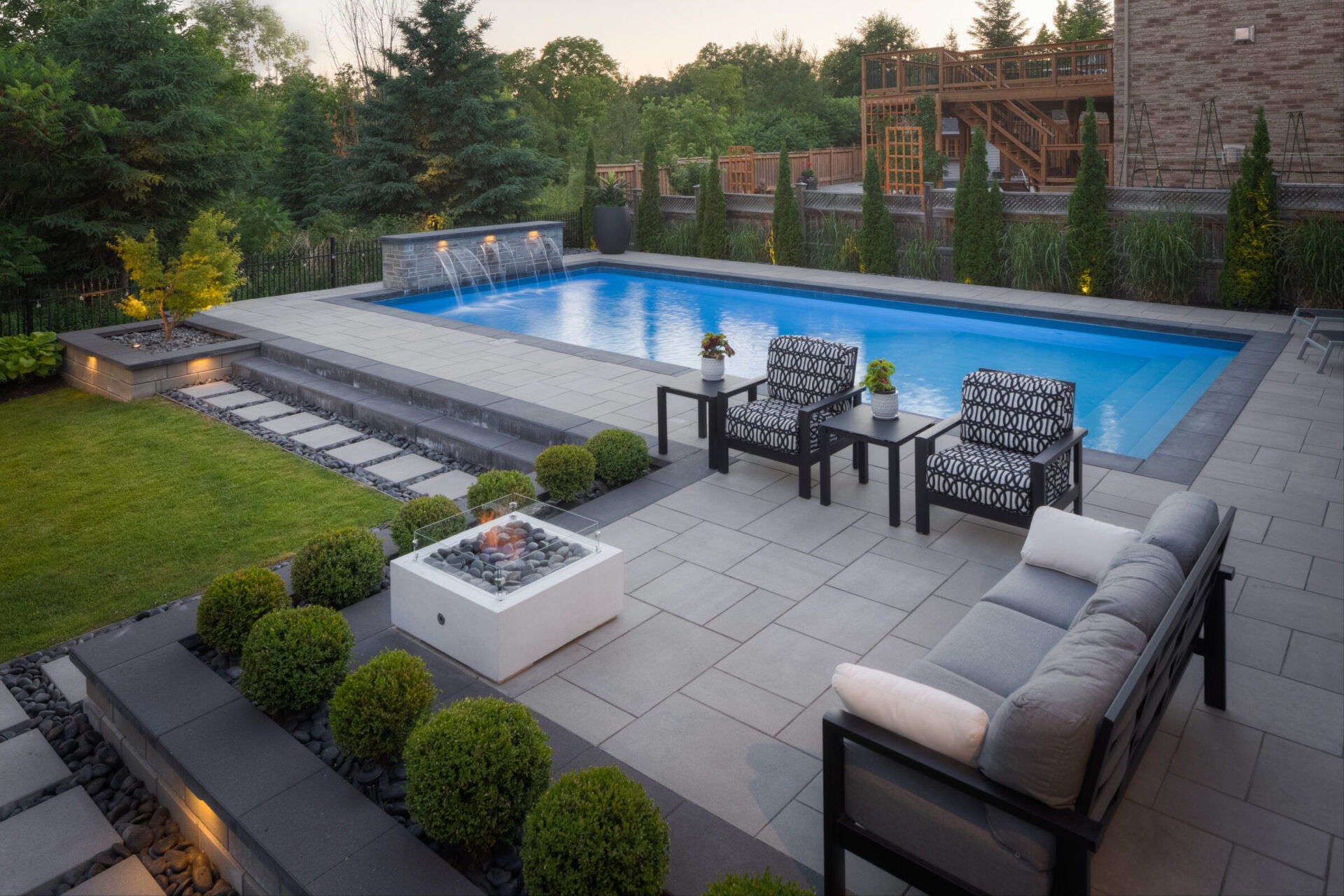 An elegant outdoor area at dusk featuring a large swimming pool, patio furniture, a gas fire pit, well-manicured lawn and landscaping, and a wooden deck.