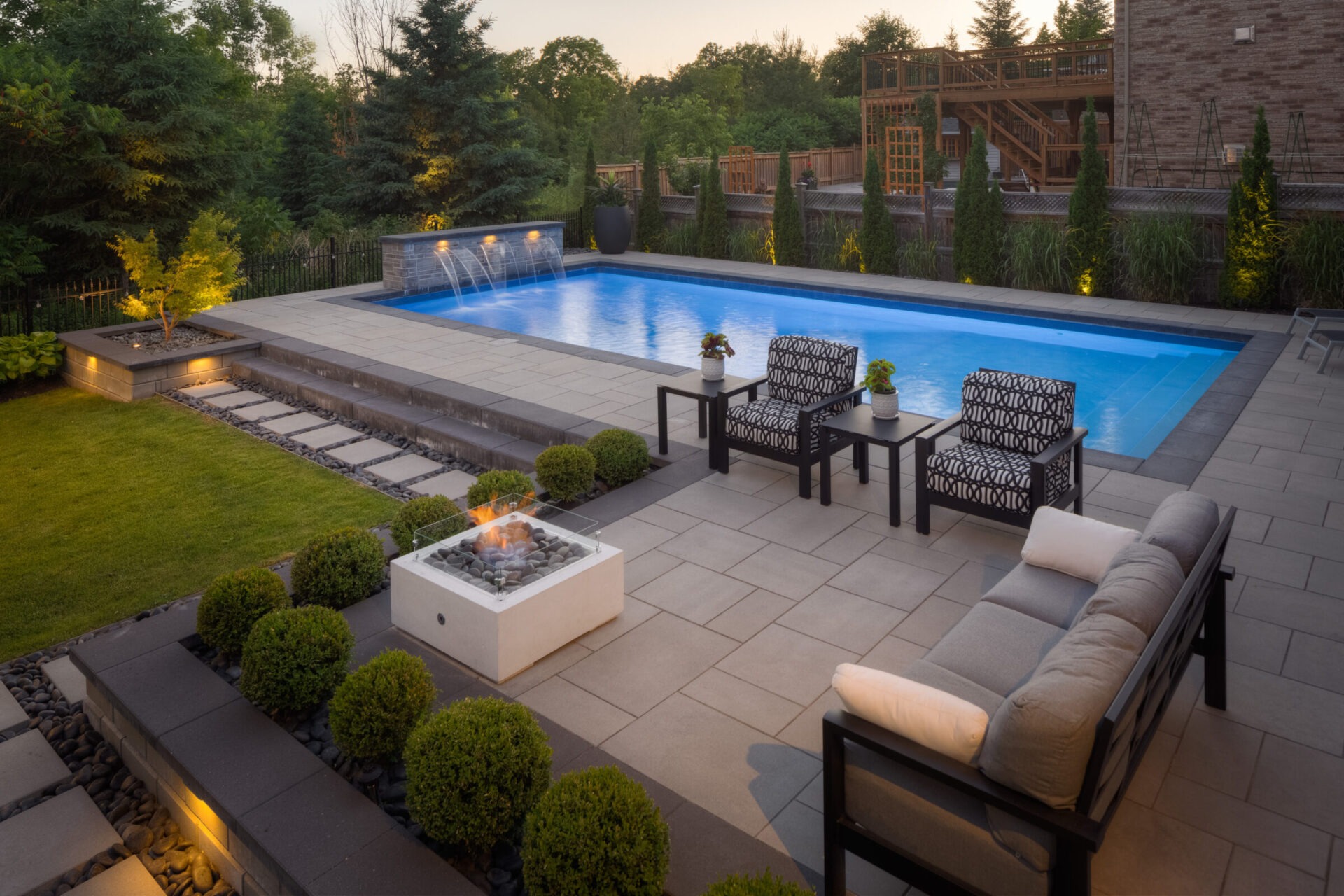 An elegant backyard at dusk featuring a lit swimming pool, patio with outdoor furniture, fire feature, manicured lawn, and ambient garden lighting.